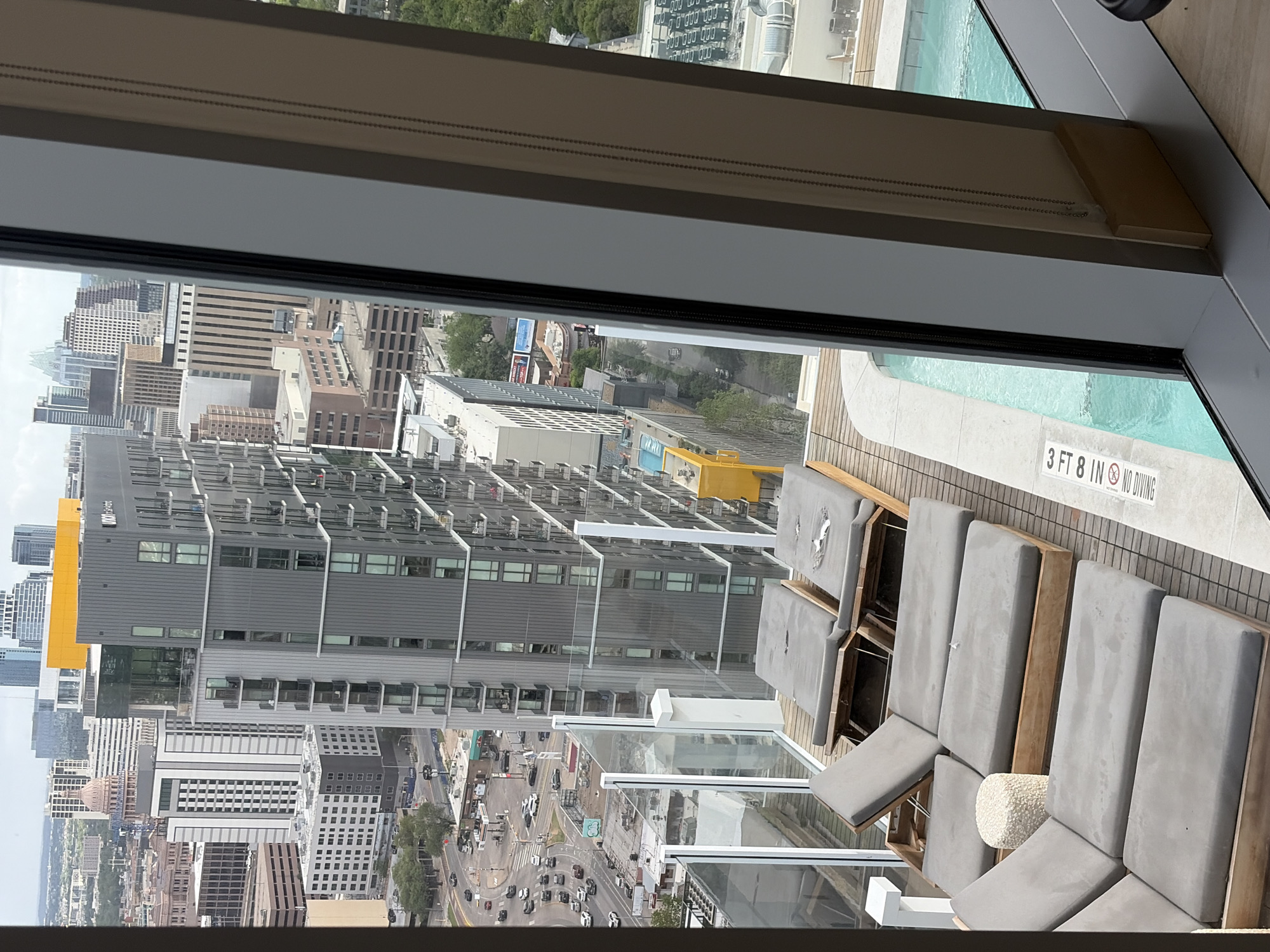 The image shows a view from a high-rise building in Austin, Texas, United States. Through a window, a cityscape unfolds, including other tall buildings, streets with traffic, and a partial view of the Texas State Capitol. Adjacent to the window, there is a pool area with a row of lounge chairs and a sign that reads "3 ft 8 in No Diving." The scene is viewed from an elevated perspective, capturing both the indoor and outdoor elements of the location.