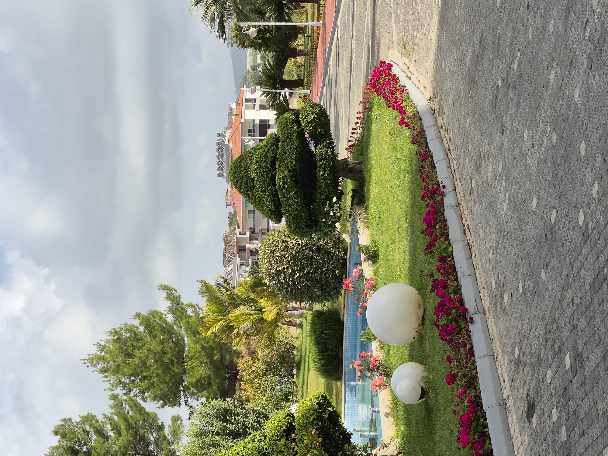 A picturesque view from Alanya, Turkey, captures a meticulously landscaped garden alongside a brick-paved pathway. The garden features topiary shrubs sculpted into conical shapes, a spherical shrub, and flower beds bursting with red blooms. A tranquil water feature, with two white spherical lights along its side, cascades a blue hue amidst the lush greenery. Buildings rise in the backdrop against a sky veiled with overcast clouds. The scene is devoid of people, focusing instead on the serene beauty of the maintained landscape.