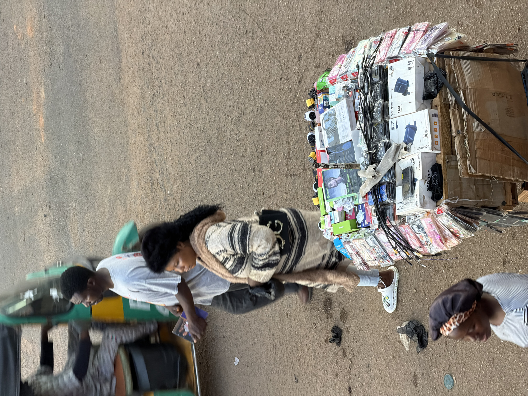 In Bukuru, Nigeria, a street scene unfolds with an abundance of items displayed for sale on a makeshift stall. The stall is a cluttered arrangement of various electronic accessories and packaged goods, primarily chargers, cables, and what appear to be phone accessories, stacked high on a wooden platform. Among the items are visible brands like "GODFREY" and "SONY," indicating electronic goods. A man in a light grey t-shirt and dark pants stands near the stall, holding a folded magazine or brochure. A woman, wearing a striped sweater and a tan scarf, stands beside him, carrying a black handbag with a prominent "GG" logo. In the foreground, a young girl with a headscarf is partially visible, looking to the side. To the left of the scene, a blurred green and yellow auto-rickshaw, a common mode of transport in Nigeria, is in motion, with a person seated inside. The ground is a rough, paved surface, typical of a street in Bukuru, with some scattered debris. The overall impression is a vibrant, bustling street market environment.
