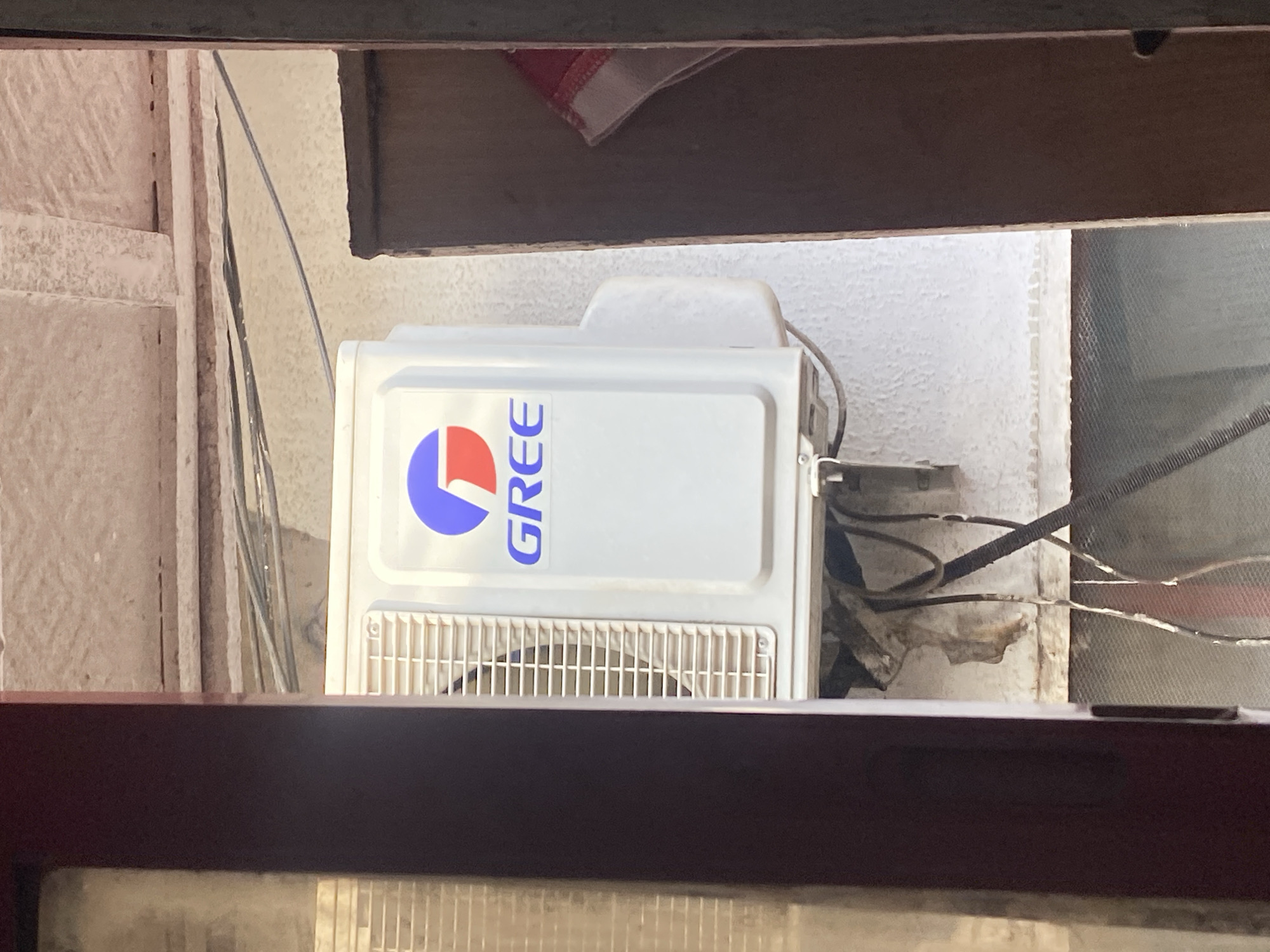An outdoor air conditioning unit, manufactured by Gree, is mounted on a textured wall, framed by a dark brown wooden structure and a red-brown metallic doorframe. The unit, primarily white, features a prominent Gree logo with blue and red accents above the brand name. Below the unit, electrical wires and conduits are visible, with some appearing weathered and disconnected. The background shows a rough, grey ceiling with exposed wiring and pipes. A portion of a red and white cloth is tucked into an opening to the right of the air conditioning unit. The scene appears to be located in Ikorodu, Nigeria, likely an exterior wall of a residential or commercial building.