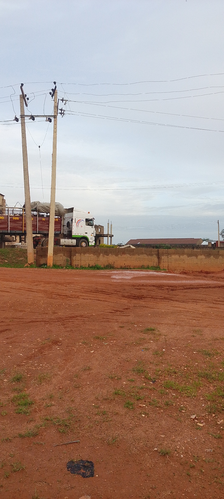 A white and green truck is parked next to a brick wall, partially obscured by power lines and utility poles. The truck is loaded with materials covered in a gray tarp. In the background, there are unfinished buildings and a roofline. The foreground consists of a dirt ground with tire tracks and some scattered grass. The sky is overcast.
