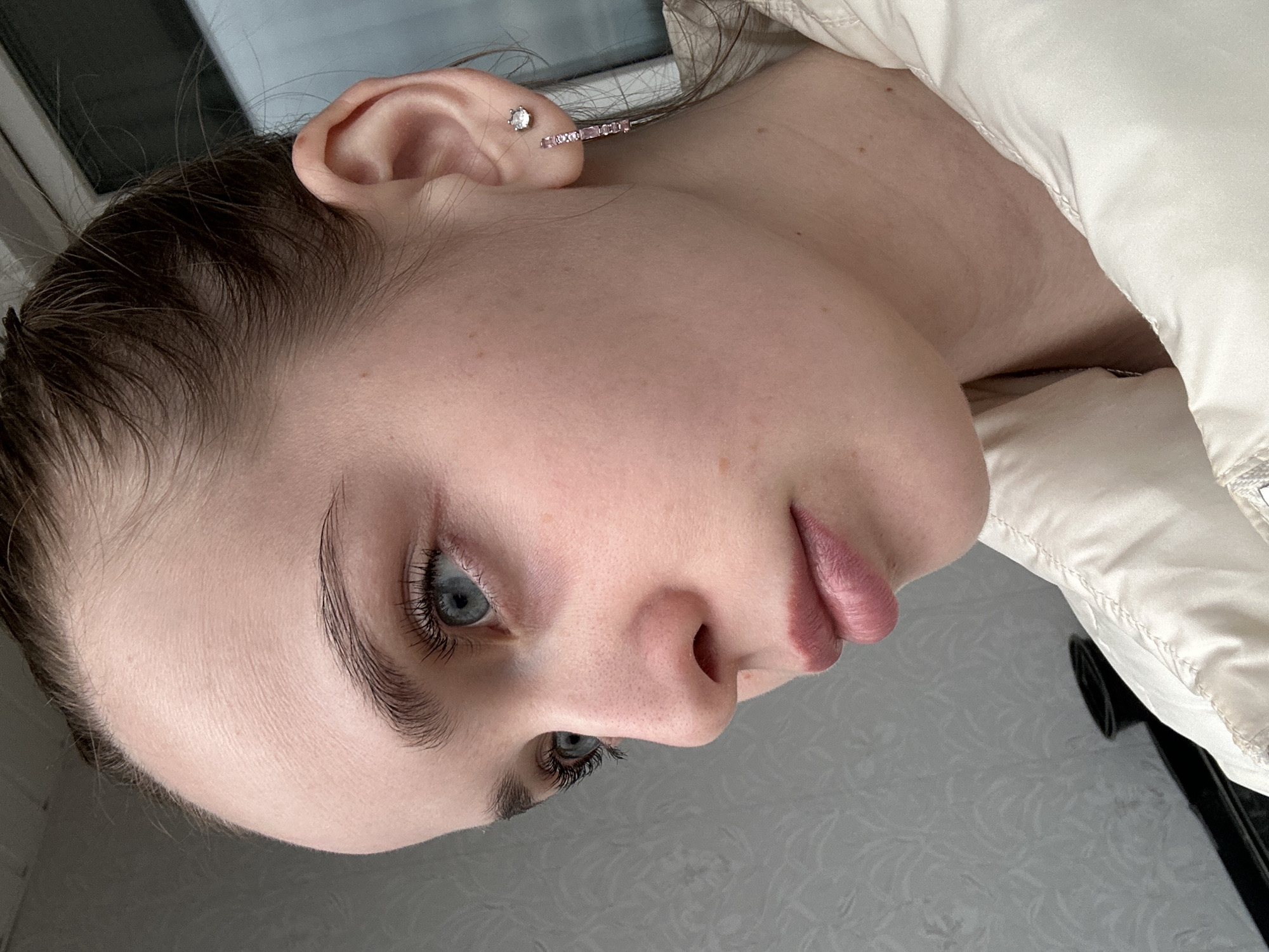 A close-up, rotated image shows a fair-skinned person with light blue eyes and dark eyebrows and hair. The person is wearing earrings, a small stud and a bar-shaped earring with pink stones, in their left ear. The person is wearing a cream-colored puffy jacket. The background includes a wall with a textured surface and a window frame in the upper left. There is nothing in the image to indicate the location is Chaikovsky, Russia.