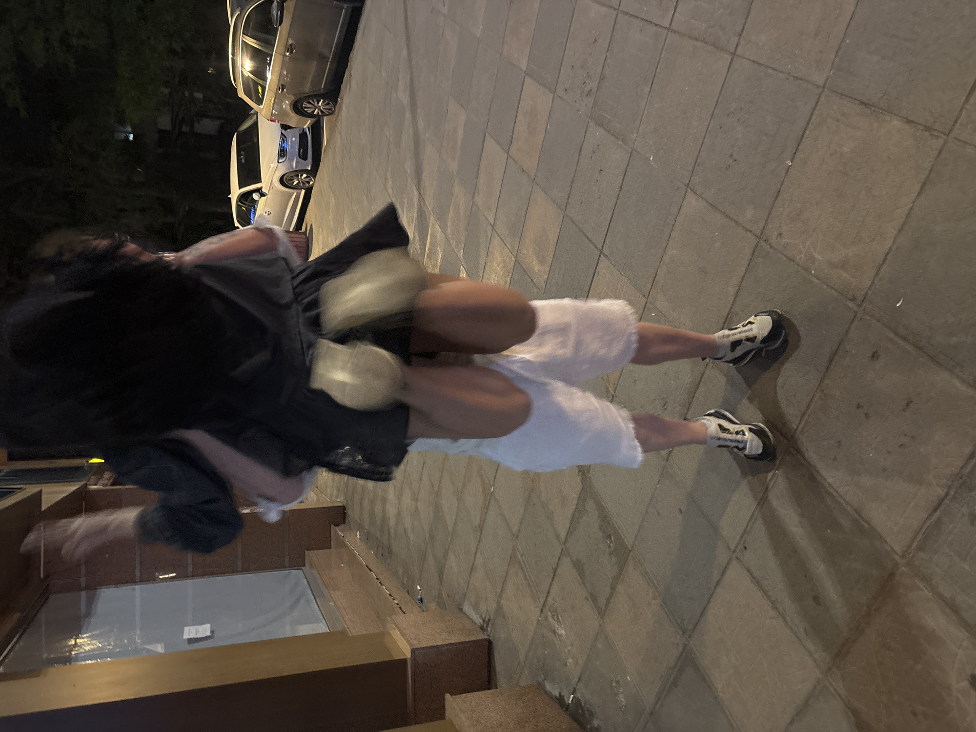A blurry image, tilted approximately 45 degrees, captures a person in motion on a brick-paved street in what appears to be an urban environment. The individual is wearing white shorts, a dark-colored jacket, and athletic shoes, and seems to be walking towards a building. A storefront with a window is visible to the left. Several cars are parked along the street in the background, suggesting a city setting. Insufficient data exists to confirm the location in Plovdiv, Bulgaria.