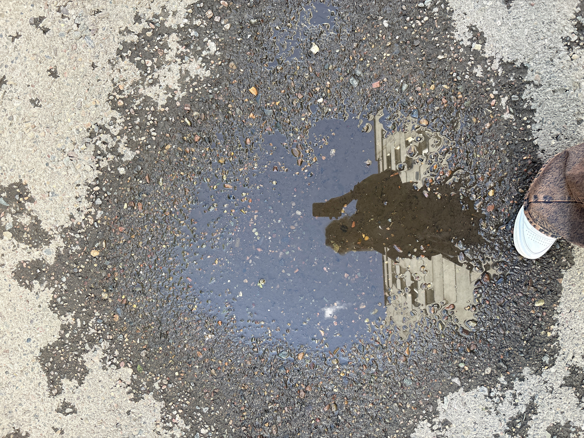 The image shows a puddle on asphalt, likely in a public space. The pavement surrounding the puddle transitions from a lighter, rougher texture to a darker, smoother texture. The puddle reflects a building with vertical lines, possibly windows or structural elements. The edge of a person's shoe, possibly a sneaker with a white sole, is visible on the right side of the puddle.
The location, Krasnoyarsk, Russia, is not directly ascertainable from the visual information.