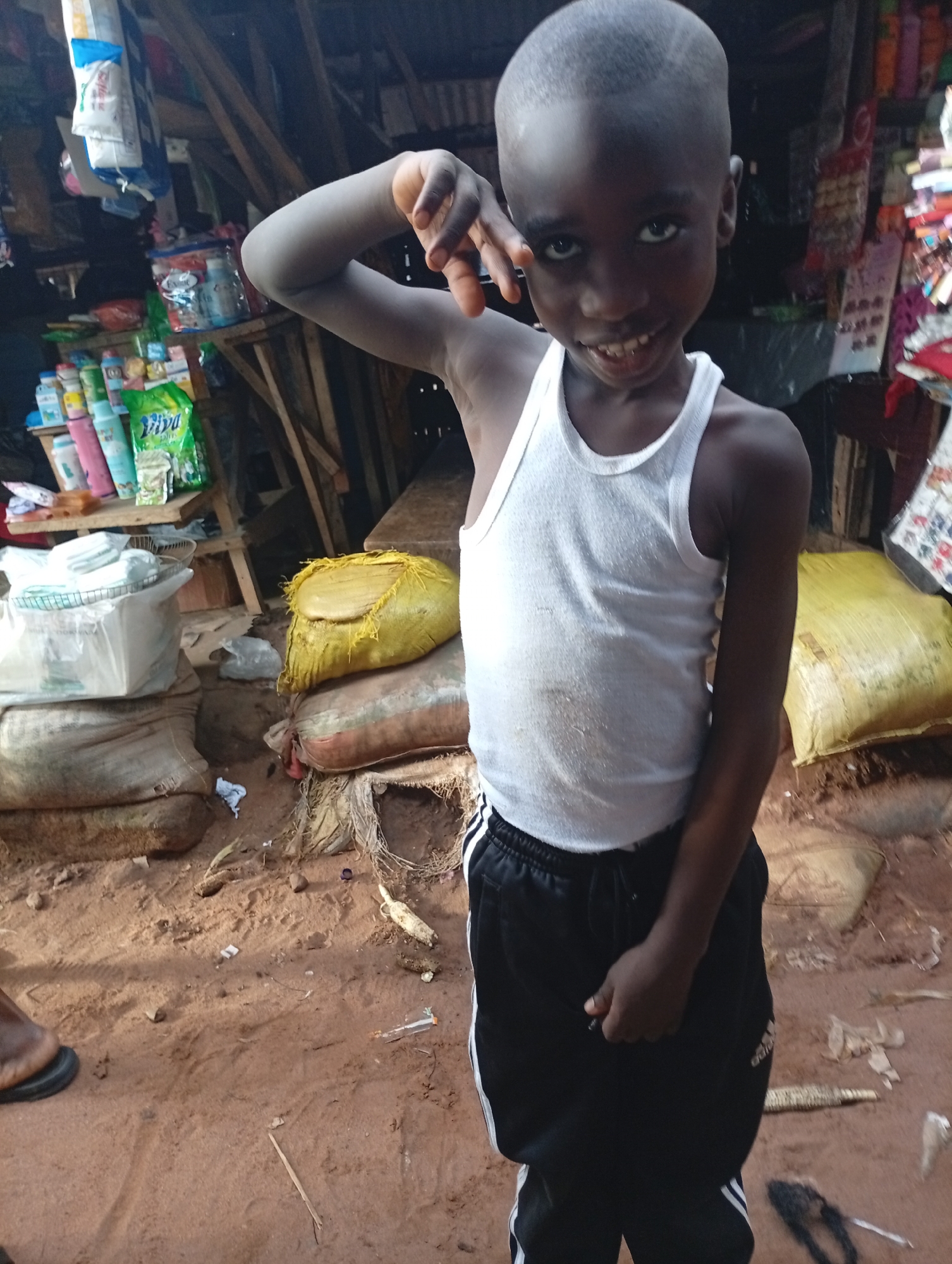 A young boy, appearing to be of African descent and around 6-8 years old, stands in what seems to be an outdoor market stall in Nkpor, Nigeria. He is wearing a white tank top and black athletic pants with white stripes. The boy is looking at the camera with a slight smile, his right hand raised to his forehead. Behind him, shelves are filled with various products, including bottles and packaged goods. The ground appears to be made of red soil, with some sacks and debris scattered around. The environment suggests a humble, possibly rural or semi-urban, commercial setting.