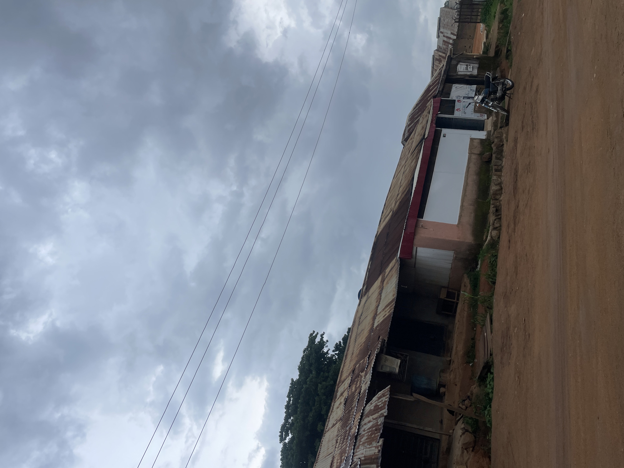 The image depicts a street scene in Gwong, Nigeria, under a cloudy sky. The foreground is a dirt road with red-brown soil. To the right of the road, a row of buildings with corrugated metal roofs are visible. These buildings appear to be shops or small businesses, with some showing signs of age and wear. One building has a pink facade with a white section. A motorcycle is parked in front of one of the shops. Behind the main row of buildings, more structures and a fence can be seen. To the left of the buildings, a large green tree stands. Overhead, power lines stretch across the frame. The overall impression is one of a typical urban or semi-urban environment in Nigeria, with a slightly overcast sky suggesting the possibility of rain.