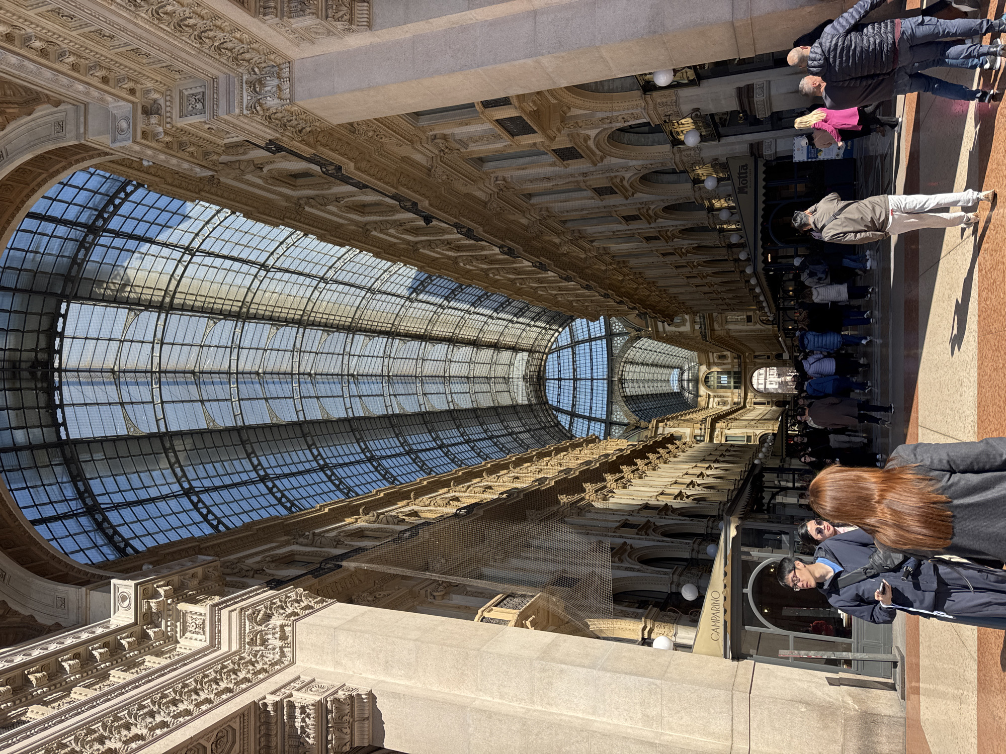The image depicts the interior of the Galleria Vittorio Emanuele II in Milan, Italy, viewed from one of its entrances, looking down the main arcade.
**Environment:**
The scene is characterized by the monumental 19th-century architecture of the Galleria. A vast, arched glass and iron roof extends overhead, allowing bright natural light to illuminate the space and showing a clear blue sky. On both sides, four-story buildings with highly ornate classical facades line the arcade. These facades feature extensive decorative carvings, cornices, balustrades, arched windows, and balconies with wrought-iron railings. Some sections of the building's exterior show protective netting. The floor is paved with a distinctive geometric pattern of light and dark tiles. Commercial establishments are visible along the ground floor, including "Camparino" and "Motta," identified by awnings and signage.
**People:**
Approximately two dozen individuals are visible in the foreground and midground, populating the wide arcade. They appear to be pedestrians or tourists, generally walking or standing in small groups.
* In the immediate foreground, facing away from the viewer, is a female with long, reddish-brown hair, wearing a dark gray ribbed jacket.
* To her left, a male with dark hair and glasses wears a dark blue jacket over a light blue shirt, dark trousers, and a black cross-body bag. He holds a white object, possibly a smartphone, in his right hand. Beside him, a female with dark hair and sunglasses is partially visible.
* Further into the arcade, a diverse crowd of people of varying ages and ethnicities are present. Most are dressed in casual attire, including jackets and long pants, suitable for a cool or bright day.
* Specific individuals include a male in a brown jacket and light trousers walking away from the viewer, and two males in the right foreground, one in a dark puffer jacket and jeans, the other in a grey t-shirt and dark jeans.
* No direct, clear interactions beyond general pedestrian flow are distinctly discernible. Many individuals are observing their surroundings or moving through the space.
**Objects:**
Beyond the architectural elements, visible objects include signage for businesses, streetlights with spherical white globes mounted on columns, and personal items carried by individuals such as phones and bags.