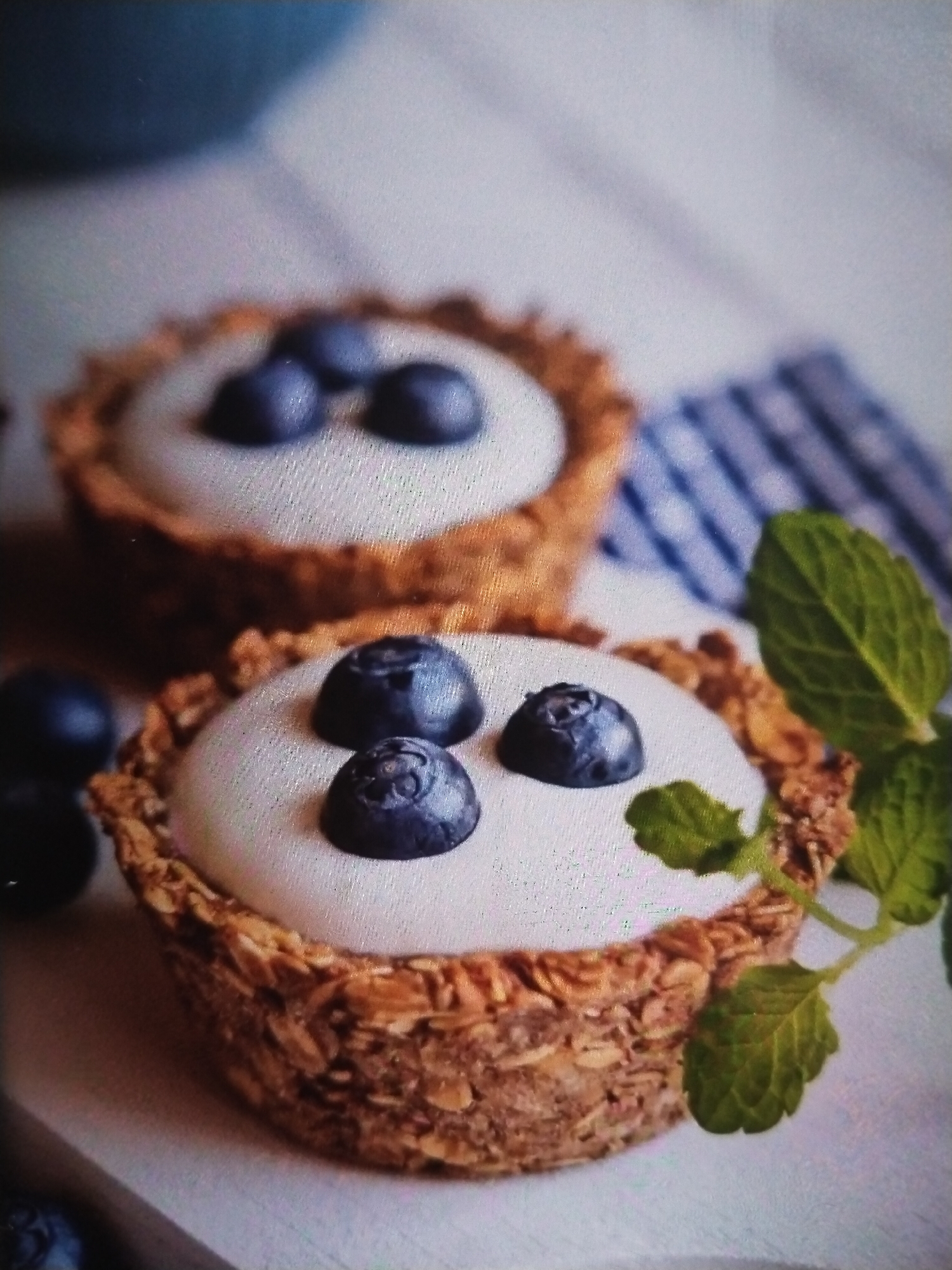 The image displays two small, individual tarts topped with a white creamy filling and fresh blueberries. The tart shells appear to be made from granola or oats, giving them a textured, golden-brown appearance. One tart is slightly out of focus in the foreground, showcasing three plump blueberries and a couple of fresh mint leaves on its right side. The second tart, positioned behind and to the left of the first, is also topped with blueberries, with three visible on its surface. The setting is a light-colored surface, possibly a table or countertop, with a blue and white striped cloth visible in the background. The lighting suggests it could be daytime, creating soft shadows and highlighting the textures of the food. No people are visible in the image. The focus is entirely on the visually appealing dessert. The overall impression is one of a light, fresh, and healthy sweet treat.