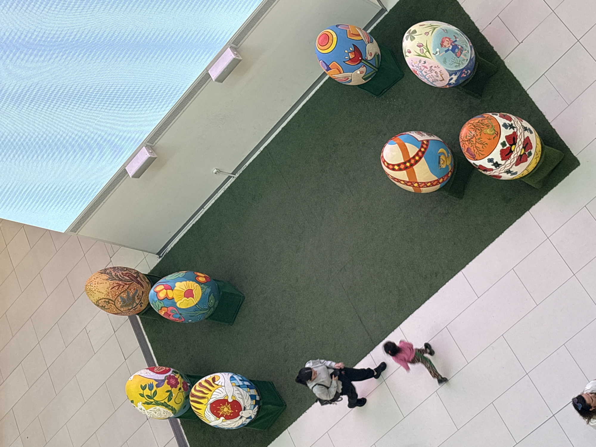 The image, taken from an overhead perspective, depicts a scene inside what appears to be a shopping mall in Plovdiv, Bulgaria. The floor is tiled with large, light-colored tiles. A rectangular area covered with green material, resembling artificial grass or carpet, is present. Scattered across this green area are eight large, decorated eggs placed on small, green stands. Each egg features a unique design.
Two adults and a child are present on the tiled floor near the green area. One adult is walking with the child, while the other adult is taking pictures. The lighting is bright, indicating an indoor environment with ample artificial light, as evidenced by the large illuminated ceiling panel.
