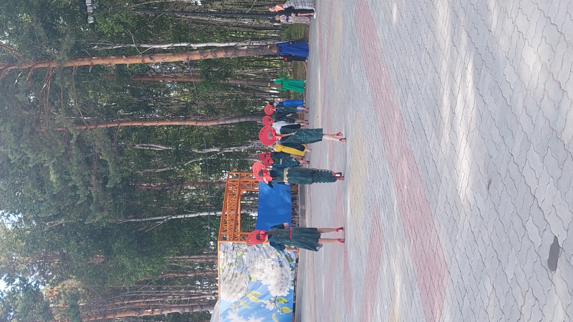 An outdoor event is taking place in Komsomolsk-on-Amur, Russia, on a paved area with a patterned surface. In the background, a line of tall trees, primarily pines and birches, forms a natural backdrop. An orange metal archway structure is visible, along with a large blue fabric screen and a mural depicting white flowers on a blue background. Several individuals, mostly young females, are walking away from the camera. They are dressed in matching dark green outfits adorned with intricate patterns and bright red, flower-shaped headwear. They are also wearing high-heeled red shoes. Some individuals in the background are wearing brightly colored dresses, including green, blue, and yellow. The lighting suggests it is daytime, with sunlight casting shadows on the paved ground. The scene appears to be a performance or parade, likely a cultural or festival event.