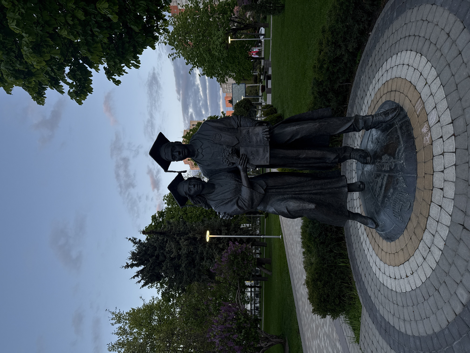 The image captures an outdoor scene at what appears to be a park or campus in Rostov-on-Don, Russia, during dusk or dawn. The central subjects are two dark bronze statues, depicting what look like graduates. Both figures wear academic gowns and mortarboard caps. The figure on the right, seemingly male, holds an open book or document in their hands. Visible Cyrillic text on this book includes "АТТ ДИПЛОМ" (likely "аттестат диплом" meaning "certificate diploma") and another word below, possibly "РЕКТОРУ" (to the rector). The figure on the left, appearing female, also holds something, possibly a bouquet or another book/scroll.
The statues stand on a circular base made of patterned pavers in concentric rings of varying shades of grey and beige, which radiates outwards. Some indiscernible chalk markings are visible on the pavement near the base. Lush green grass and dark green bushes surround the paved area. Several trees, both deciduous and evergreen, frame the scene, with dense foliage visible in the foreground and background. In the middle distance, a tall park lamp with an illuminated head is visible, and further back, the outlines of urban buildings can be seen against the sky. The sky itself is a soft blue-grey, with scattered clouds showing faint hints of pink or orange, consistent with twilight hours. No active event or living people are depicted; the scene is static, focusing on the monument.