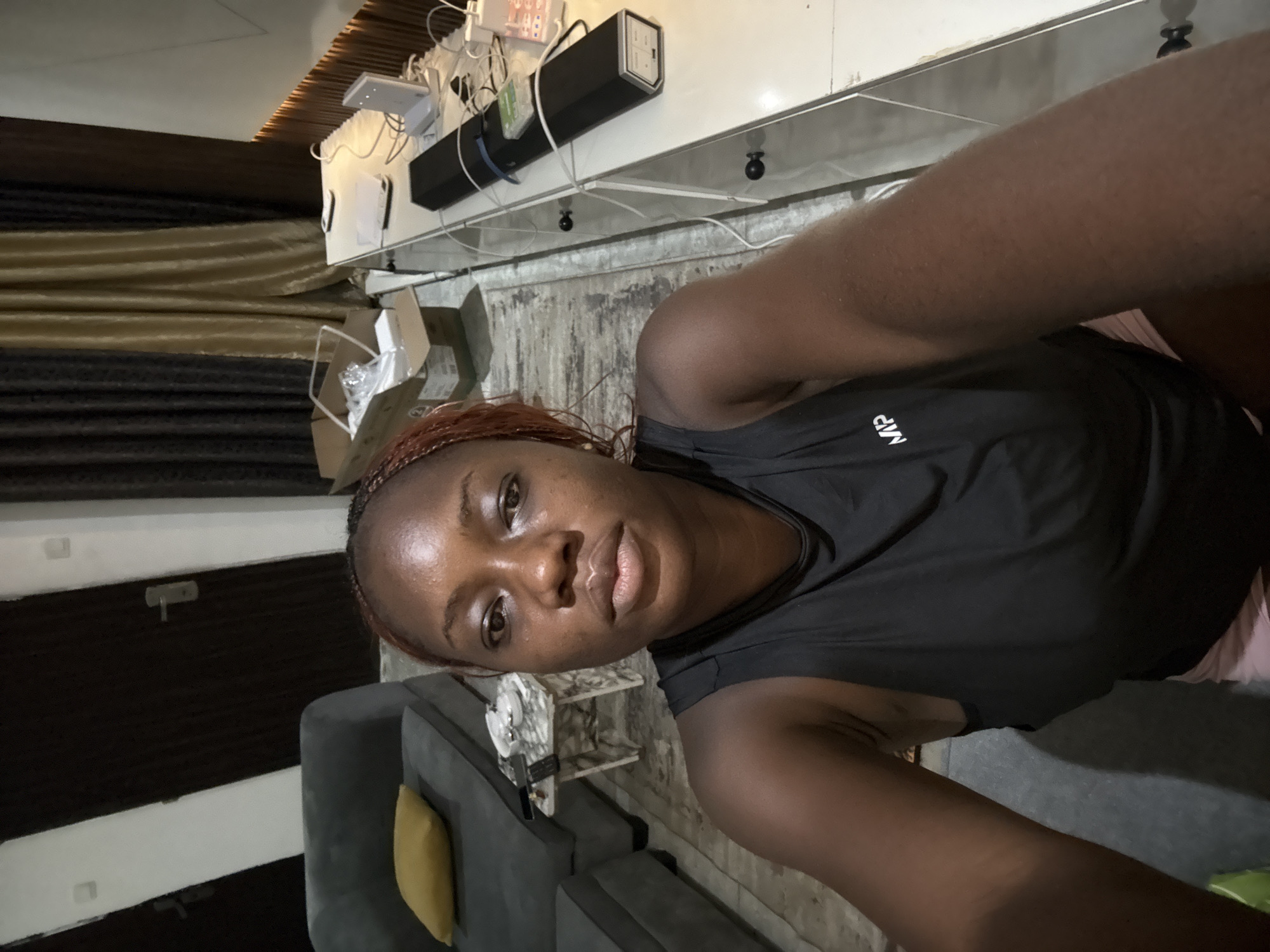 A medium-shot, self-portrait photograph presents a dark-skinned woman wearing a black tank top with the letters "MP" visible on the chest. Her hair is reddish-brown and braided. The woman is indoors, as evidenced by the interior background. A grey sofa with a yellow throw pillow is visible in the lower left corner, and a small table with what appears to be a marble top is placed next to it. Behind the woman, along the top of the frame, there is a white surface with various electrical devices and cables visible, suggesting it might be a shelf or a table. There's also a section of a curtain with gold stripes. The location is Iddo Tudunwada, Nigeria.