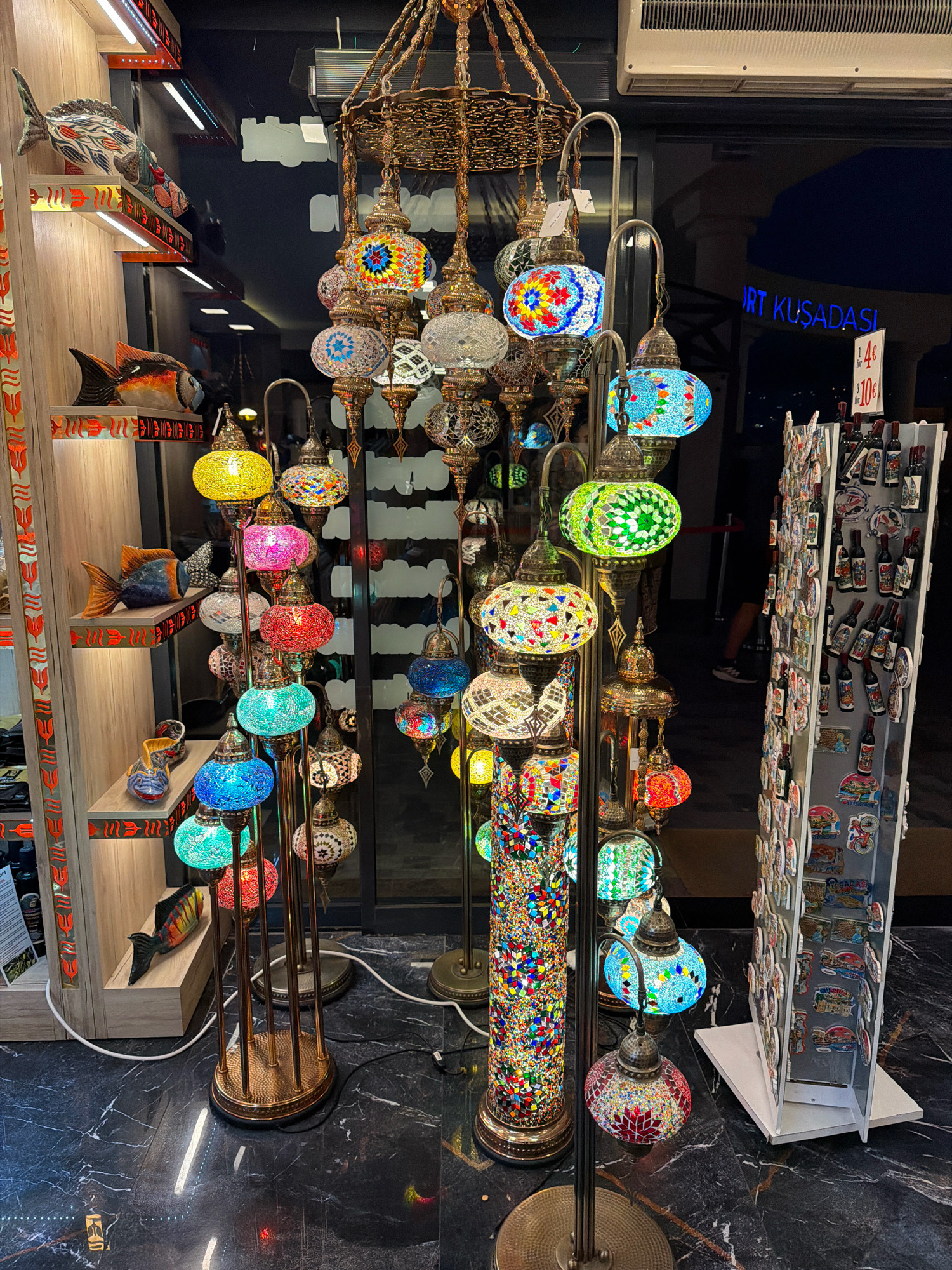 The image shows a store in Kuşadası, Turkey, displaying a variety of mosaic lamps. The lamps are of various sizes and colors, with intricate patterns. They are hung from the ceiling and displayed on stands. Some of the lamps are lit, casting a warm glow on the surroundings. In the background, there is a large window with a reflection of the store's interior. The store appears to be a souvenir shop, as indicated by the presence of postcards and other small items on display. The floor is covered with black marble tiles. There are no people visible in the image.