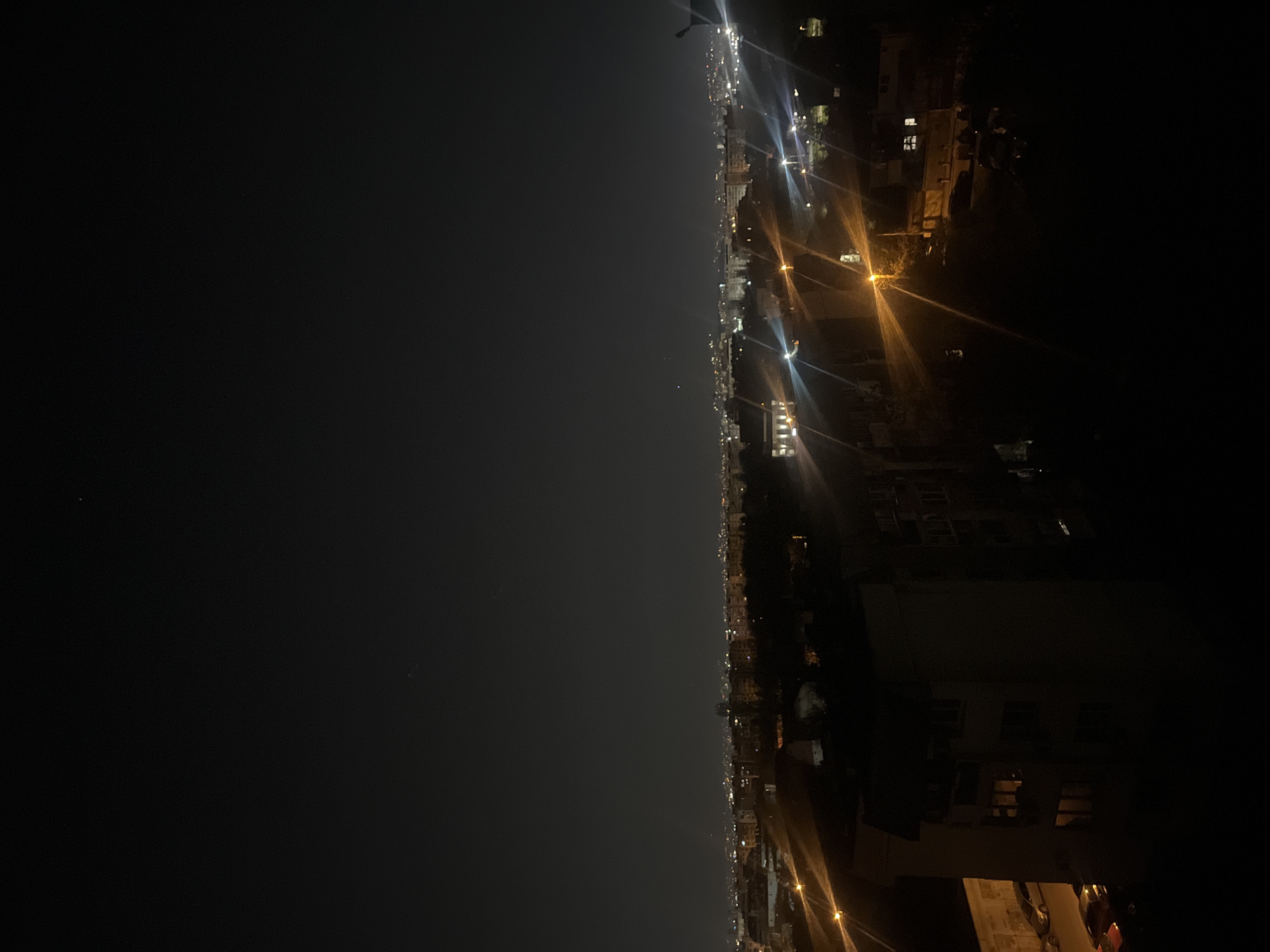 This image captures a vertical, expansive night view of an urban landscape, likely Sofia, Bulgaria, from a high vantage point. The scene is bisected by the city's horizon line, separating the dark night sky from the illuminated cityscape.
The upper portion of the image is dominated by a deep, dark night sky, appearing clear with no visible clouds. A few extremely faint specks of light are barely discernible, potentially distant stars or aircraft.
The lower half of the image showcases the sprawling city, characterized by a multitude of artificial light sources. Numerous streetlights, emitting both warm orange and cooler white light, create bright flares that define streets and illuminate facades of buildings. Many buildings appear as dark silhouettes against the glow, with various windows lit up, indicating ongoing residential or commercial activity. The density of lights suggests a vibrant and extensive urban area. While no specific individuals are visible, the widespread illumination implies active city life. The weather appears to be clear, allowing for good visibility of the distant city lights. There is no visible text in the image.