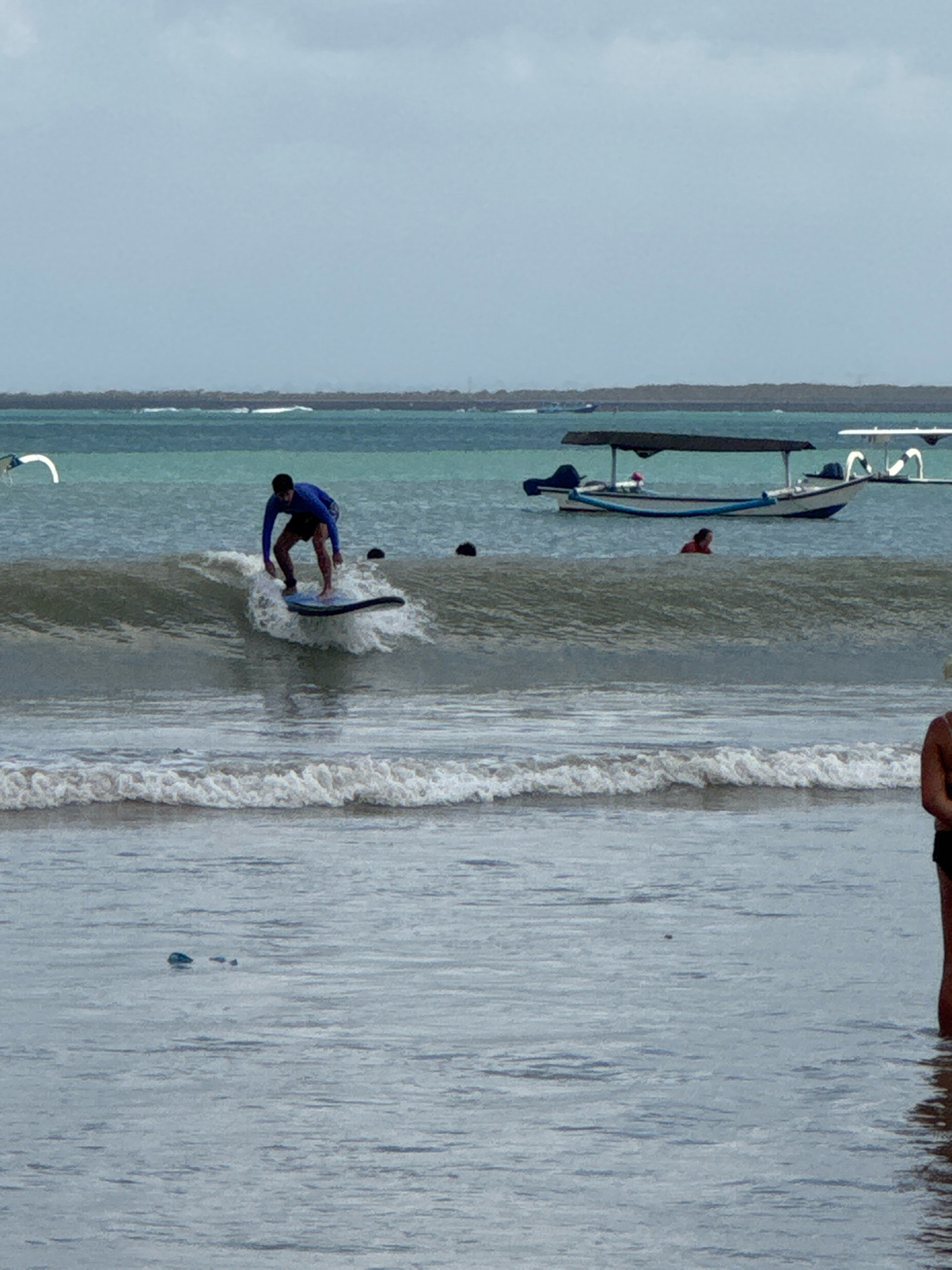 In Kuta Selatan, Indonesia, a surfer is captured mid-action on a wave in the ocean. The surfer, wearing a blue long-sleeved shirt and dark shorts, is on a light-colored surfboard, skillfully balancing as the wave crests. To the left of the surfer, a white, curved object is partially visible above the water. In the background, a boat with a dark canopy is moored, with several white poles extending from its side. Further back, a thin strip of land with vegetation lines the horizon, separating the turquoise ocean from a cloudy, pale sky. Two black buoys are visible in the water near the surfer. In the foreground, gentle waves roll towards the shore. To the right, the legs of a person standing in the water are visible, facing away from the camera.