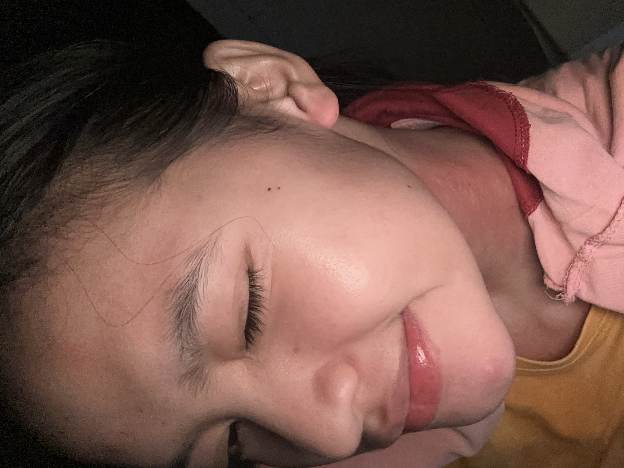 A close-up indoor shot depicts a person with dark hair lying down, facing right. The person's eyes are closed, and a mole is visible on their left temple. They are wearing a pink shirt with a maroon collar, layered over a yellow garment. The location is unknown and cannot be determined based on the image alone.