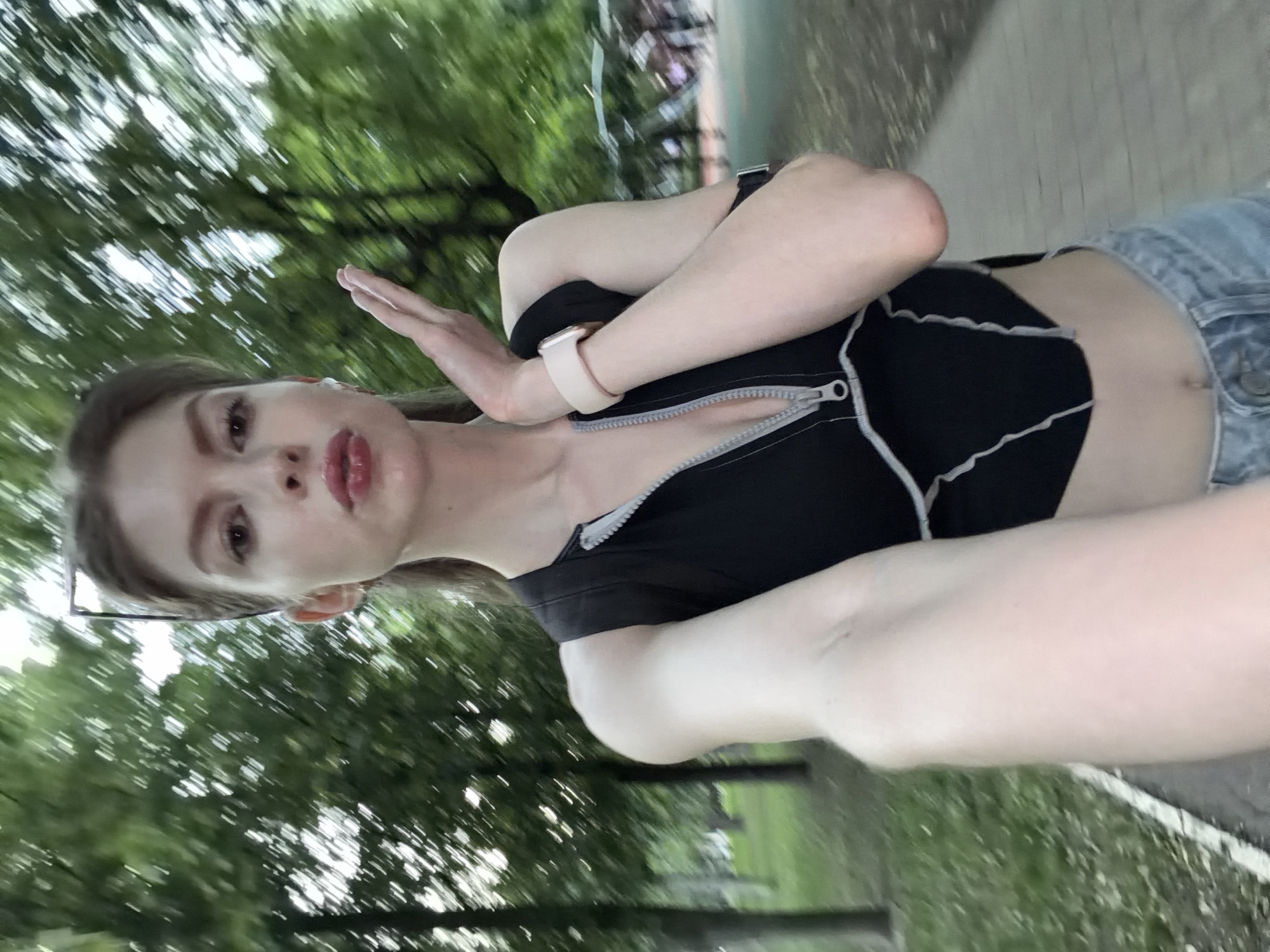 A young woman is taking a selfie outdoors. She wears a black crop top with a zipper and light-wash denim shorts. She has fair skin and is wearing a smartwatch. Her left hand is raised as if gesturing or blocking. The background shows a park environment with trees and a walkway. The photo seems to be taken in motion, giving a blurred effect to the foliage. The location is possibly a park in the city of Moscow, Russia.