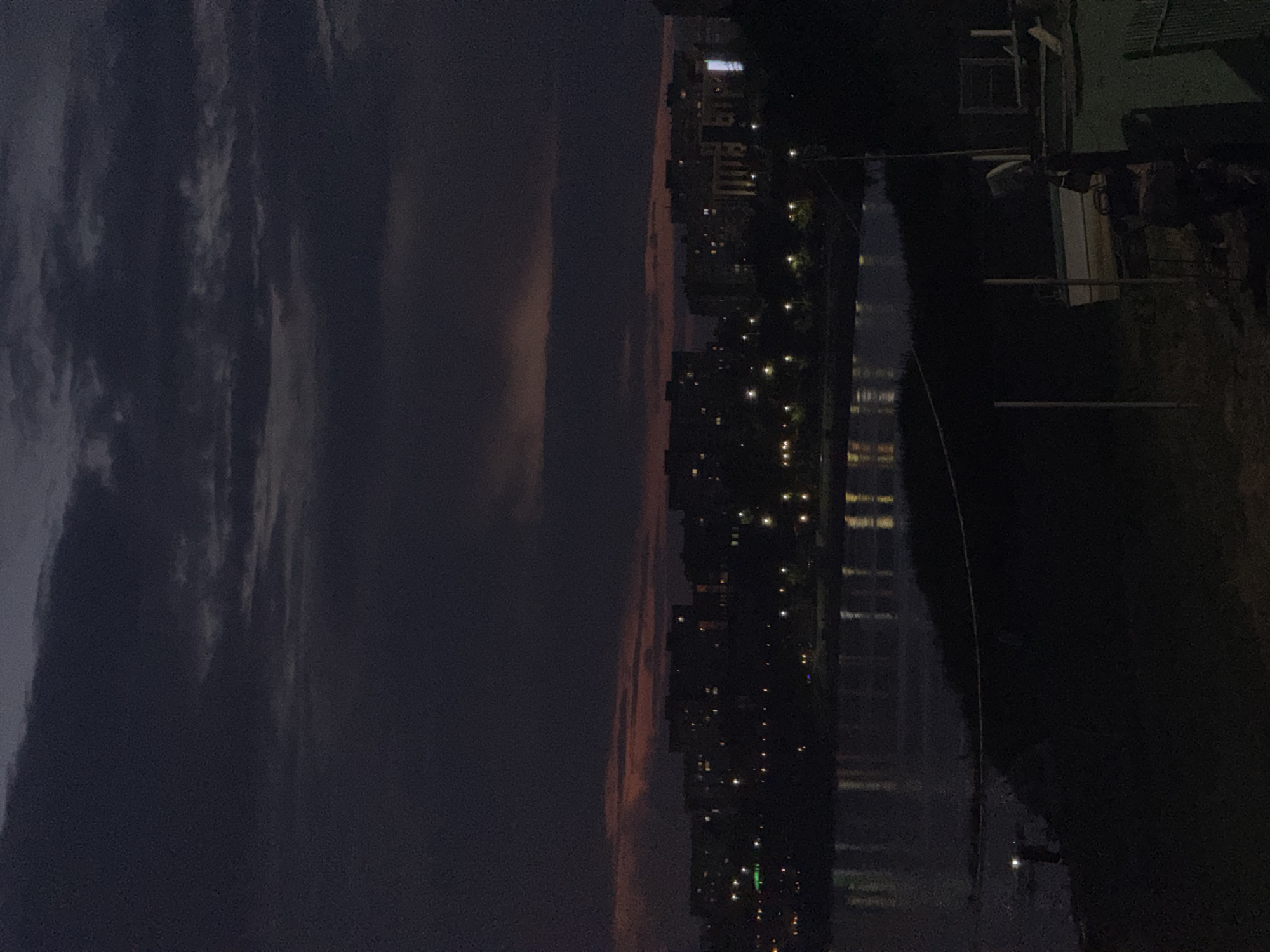 The image depicts a cityscape at twilight, with buildings and lights reflecting in a body of water. The sky is a deep purple with streaks of lighter clouds, suggesting dusk or early nightfall. A collection of tall, modern buildings are visible along the right side of the frame, illuminated by numerous windows and streetlights, creating a vibrant contrast against the darkening sky. The water in the foreground, possibly a river or canal, mirrors the cityscape, adding depth and a sense of tranquility to the scene.
On the far right, a section of a green roof and a white structure, potentially a small building or shelter, are visible, casting shadows on the ground. The overall impression is one of urban life transitioning into night, with the lights of the city creating a dynamic and lively atmosphere. There are no visible people or specific events taking place. The location is identified as Pavlodar, Kazakhstan.