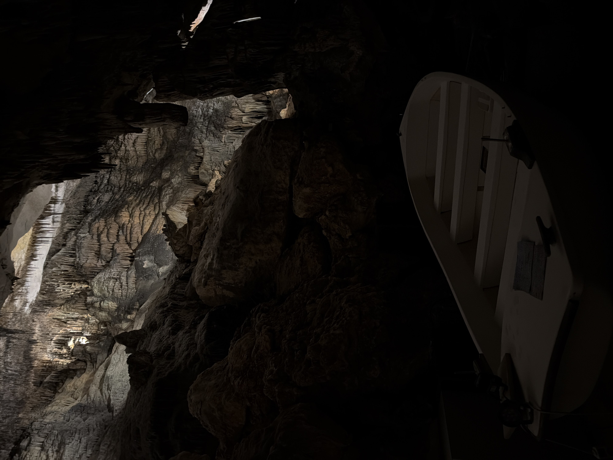The image captures a dimly lit cave interior, possibly located in Manacor, Spain. The left side features a wall formation with stalactite-like structures, illuminated by a light source. A large rock formation occupies the center of the image. To the right, a white, framed object hangs from the cave ceiling, partially obscured by darkness. The environment is dark and cavernous. There are no visible people in the scene. The overall impression is that of a natural cave formation with an added, man-made structure.