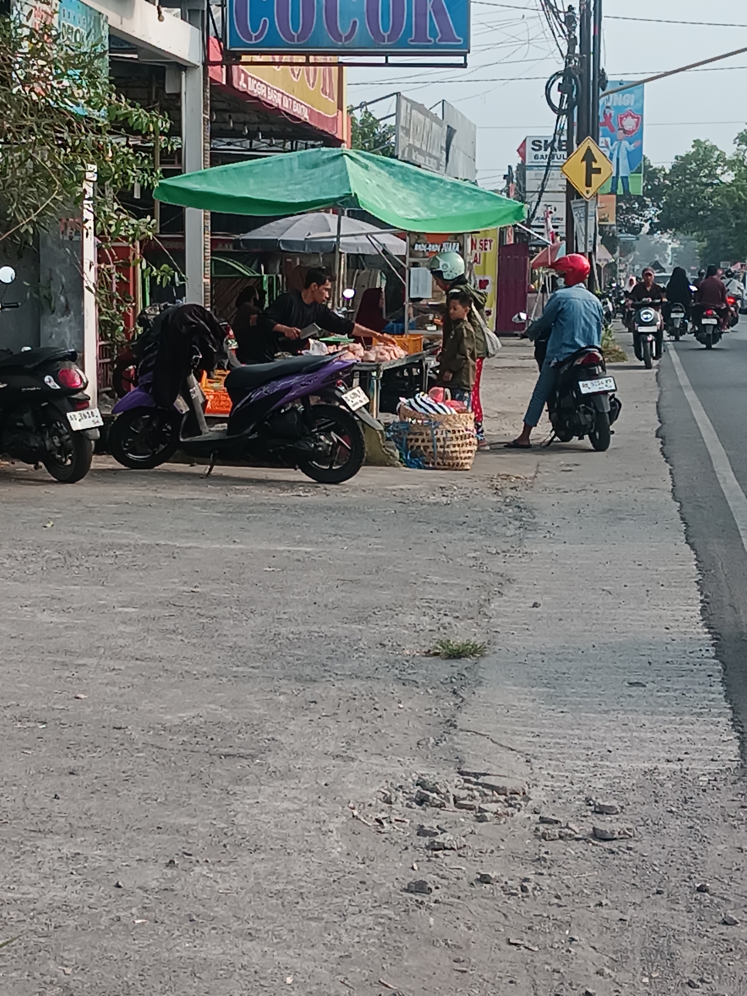This is a daytime, eye-level, medium shot capturing a street scene in Sewon, Indonesia. The foreground is dominated by a grey, cracked concrete sidewalk. In the mid-ground, a busy street is visible with several people and motorcycles. A man in a blue shirt and blue jeans, wearing a red helmet, is riding a black motorcycle heading away from the camera. To his left, a vendor stall with a green canopy is set up, featuring a man in a dark shirt and a child in a green jacket. The vendor appears to be preparing food, with baskets of goods and what looks like raw meat on display. Several parked motorcycles, including a black scooter and a purple scooter, are positioned along the sidewalk. In the background, buildings and more motorcycles on the road can be seen, suggesting a commercial or residential area. A traffic sign pointing upwards with an arrow is also visible on a pole. The overall impression is one of a typical day in a lively Indonesian town.