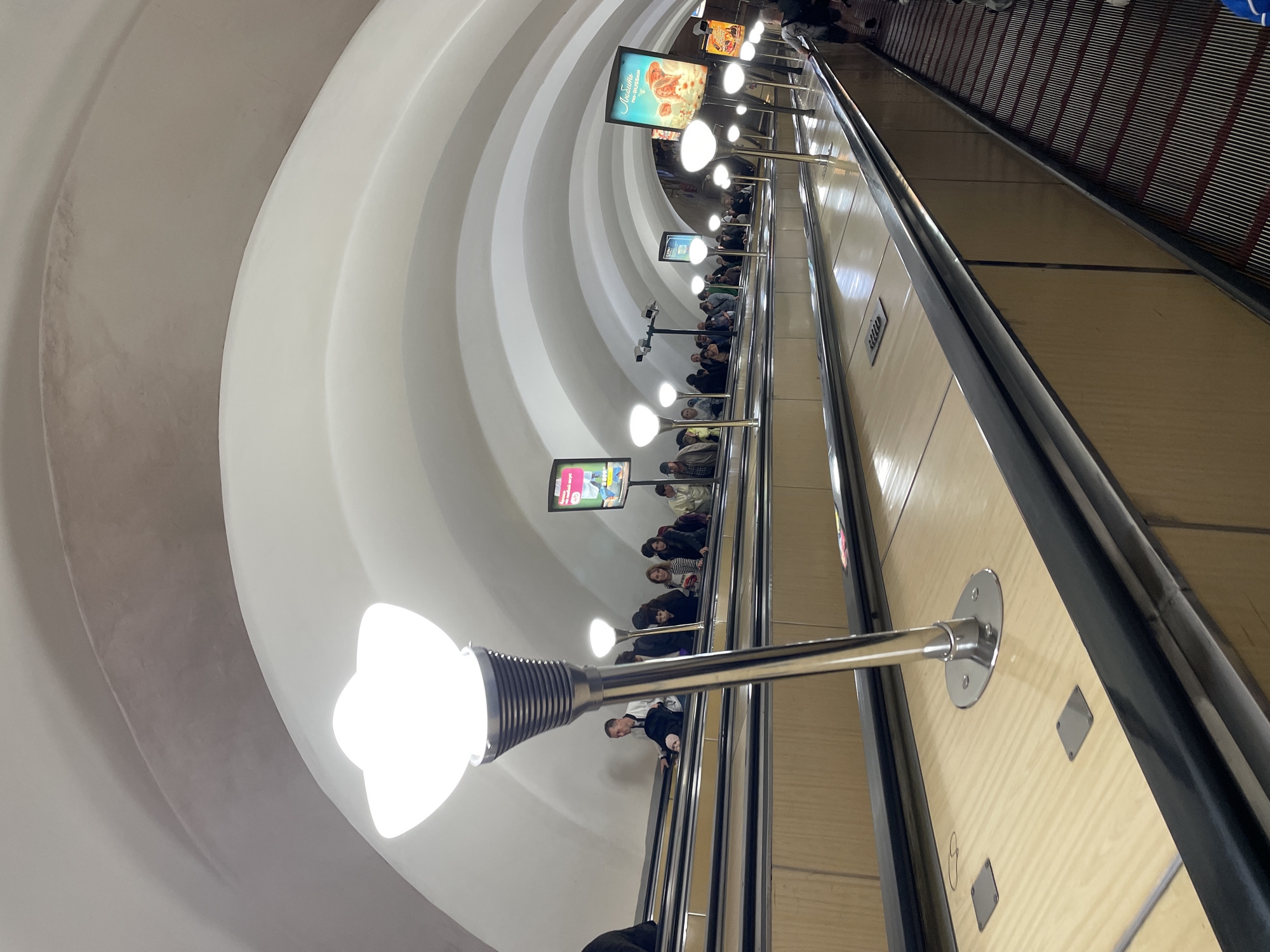 The image depicts an underground metro station in Saint Petersburg, Russia, featuring a wide, ornate escalator system. The escalator is flanked by polished, light-colored wooden panels and framed by black metal railings. Numerous people are ascending the escalator in staggered rows, facing forward, with various expressions and casual attire. The station's architecture is characterized by a series of graceful, white arched ceilings, reminiscent of a tunnel or a grand hall. Several modern, globe-shaped light fixtures are strategically placed along the sides of the escalator, illuminating the scene with a bright, white glow. Advertisements are displayed on illuminated screens mounted on the station walls, adding a splash of color to the otherwise monochromatic environment. The overall impression is one of a busy, well-lit, and architecturally significant public transportation hub.