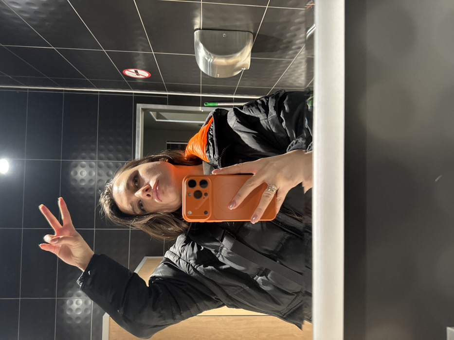 A woman takes a selfie in a bathroom mirror, holding an orange phone case. She is wearing a black puffer jacket with an orange lining. Her hair is brown and shoulder-length. She flashes a peace sign with her left hand. The bathroom has black tiled walls, a hand dryer, and a no-smoking sign on the ceiling. A doorway is visible in the background, leading to another room. The mirror reflects her image and parts of the bathroom.