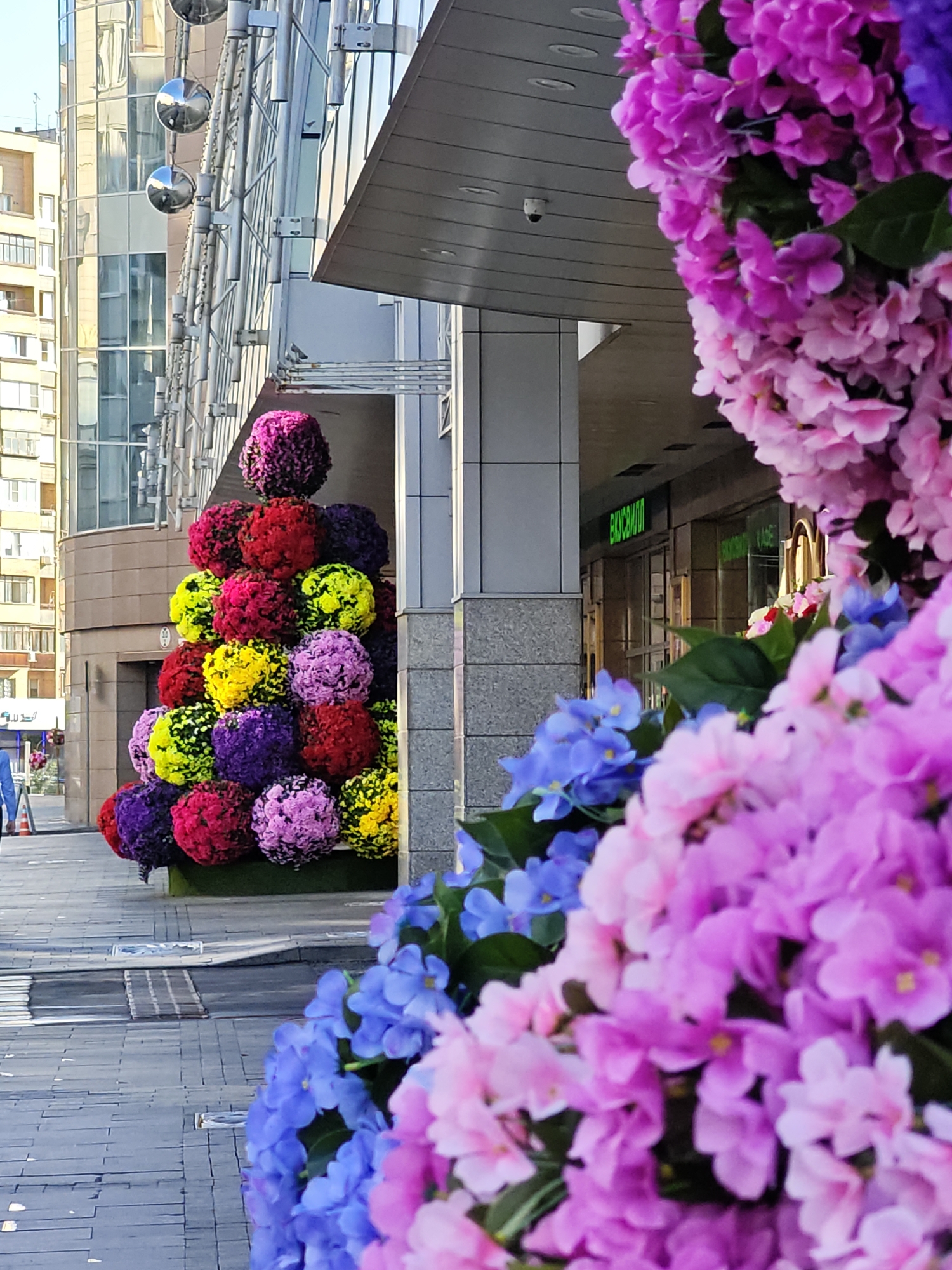 The image shows a street scene in Moscow, Russia, with a modern building facade featuring a curved glass exterior and metallic architectural elements. In the foreground, there is a large, pyramid-shaped floral arrangement composed of numerous spherical clusters of artificial flowers in vibrant colors, including pink, purple, red, yellow, and green. To the right, more artificial flowers, predominantly pink and blue, are densely clustered, obscuring parts of the building. The ground is paved with gray interlocking stones. A street sign with green lettering is visible on the building. A small security camera is mounted on the underside of an overhang. The scene is illuminated by daylight, casting shadows on the pavement.