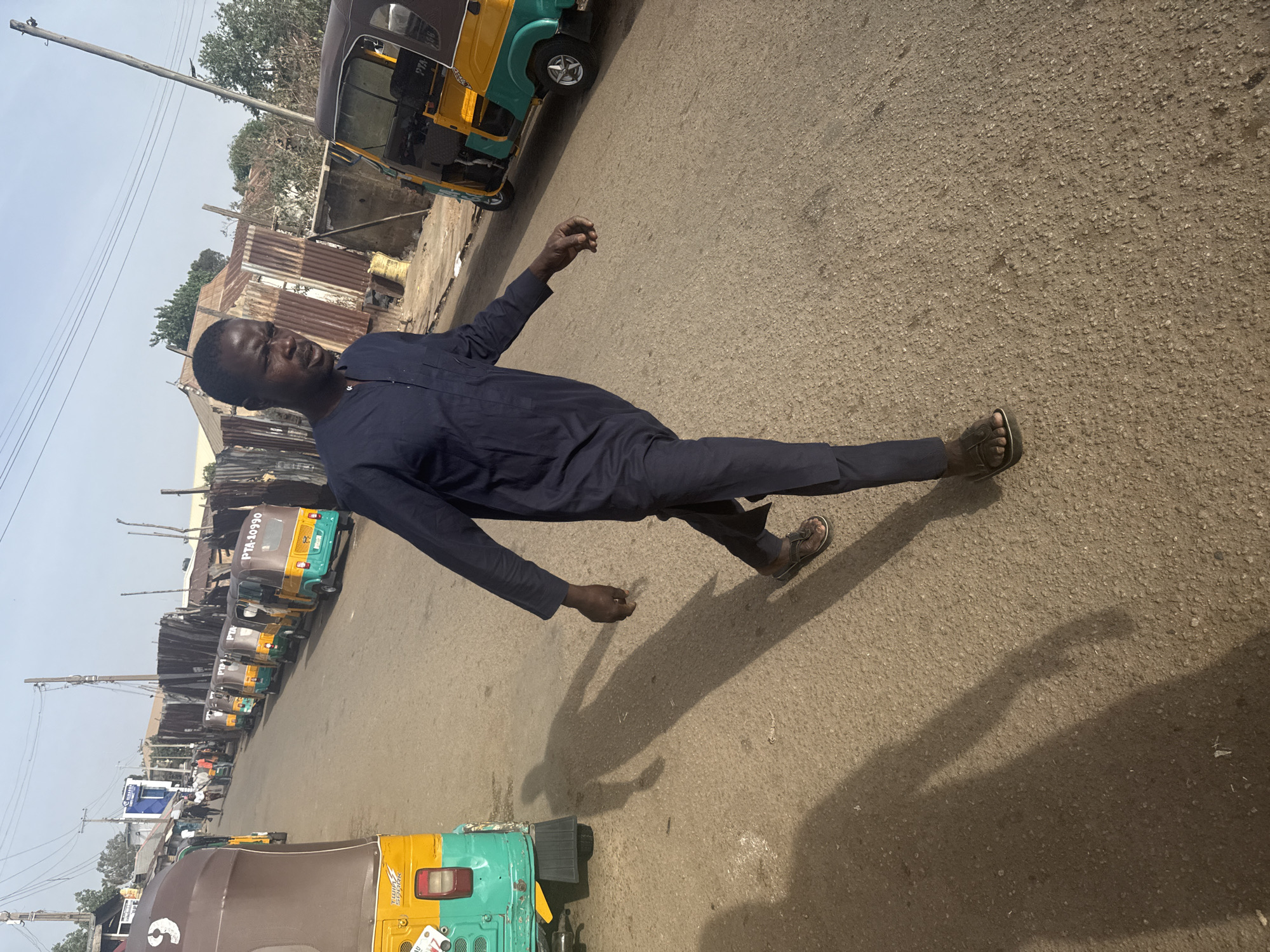 A man stands on a street in Jos, Nigeria, wearing a long-sleeved dark blue outfit and sandals. He is surrounded by several three-wheeled vehicles, locally known as "Keke Napep," painted in yellow and green. The road is paved, and buildings with corrugated iron roofs are visible in the background. The sky is overcast.