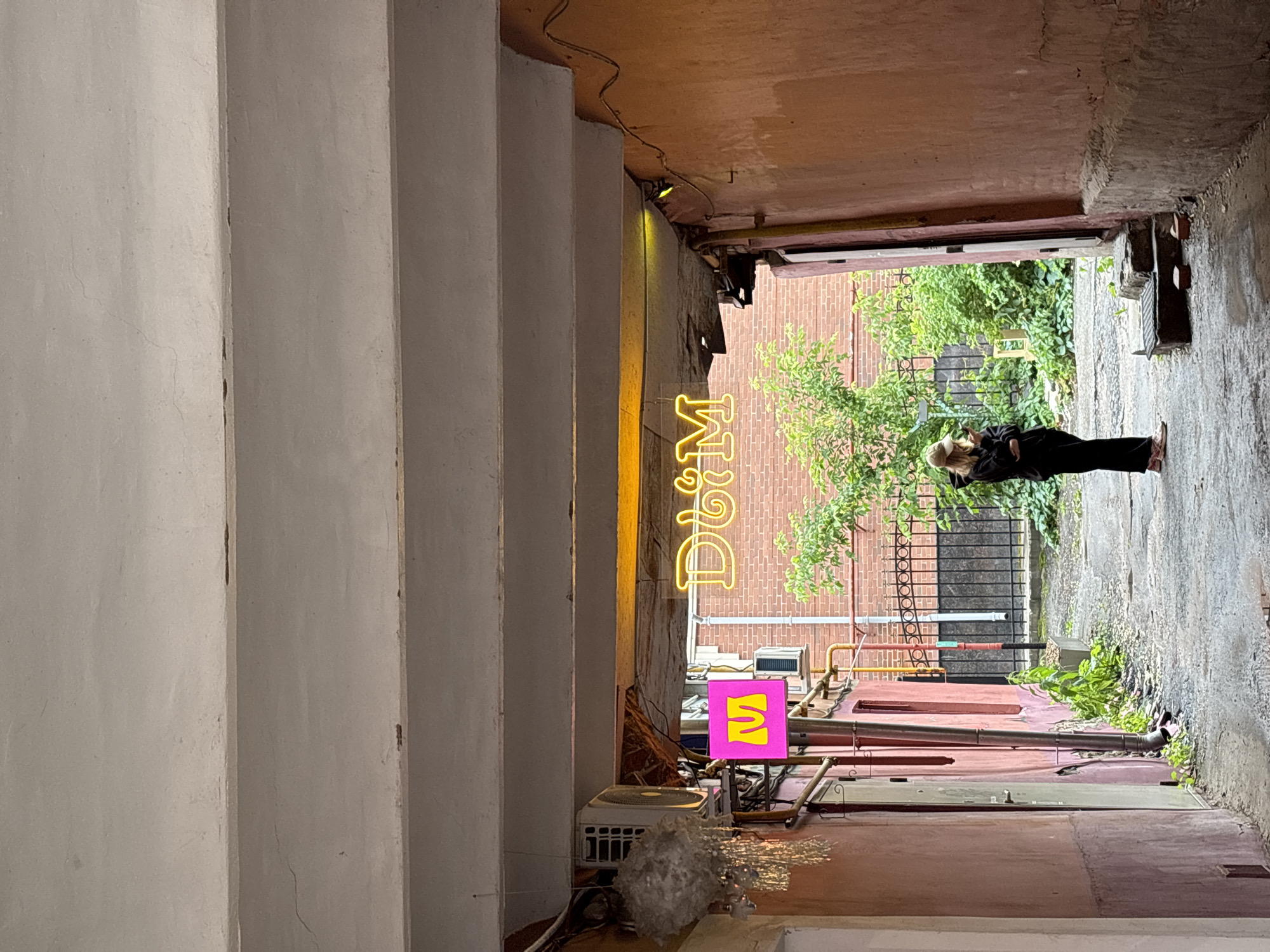The image depicts a narrow courtyard or alleyway, likely in the city of Nizhny Novgorod, Russia, as indicated by the prominent neon sign displaying "Dom" in Russian Cyrillic letters. The alley is bordered by buildings with weathered pink and red facades. A person, dressed in black attire including a hooded sweatshirt and pants, stands in the middle of the alley, looking down at their phone. They are wearing a light-colored baseball cap and light-colored sneakers with pink accents. To the left, a pink square sign with a yellow "M" is illuminated, and above it, an outdoor air conditioning unit is mounted. The ground is paved with asphalt and shows signs of wear, with puddles of water visible in some areas. Lush green foliage and a black metal fence can be seen in the background, suggesting a small garden or yard. The overall impression is one of an urban, possibly slightly run-down, but atmospheric setting.