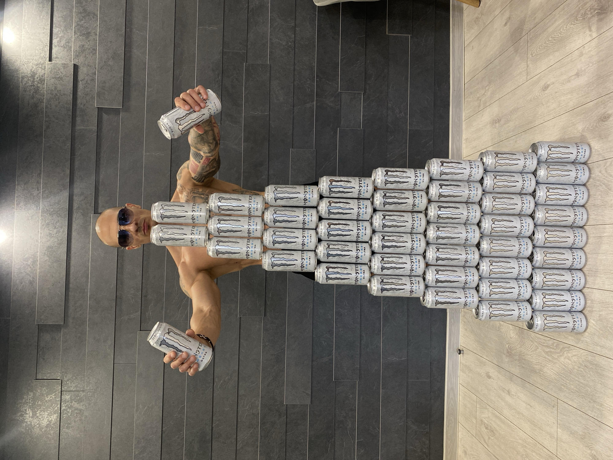 A person with a shaved head and sunglasses is lying on a floor with a tile/wooden plank design. The person is surrounded by and partially covered with numerous white Monster Energy drink cans, stacked in rows to resemble a body. The person is holding two cans aloft, one in each hand. The setting is indoors. There is no indication of location in Pleven, Bulgaria.
