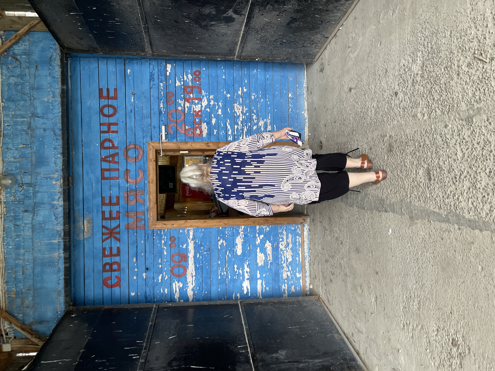 An older woman is entering a small building. The building has a blue wooden facade with peeling paint. Red Cyrillic lettering, likely a sign, is painted on the facade. The woman is wearing a patterned blouse, black pants, and brown shoes. She is carrying a small bag. The building is located in Киясово, Russia.
