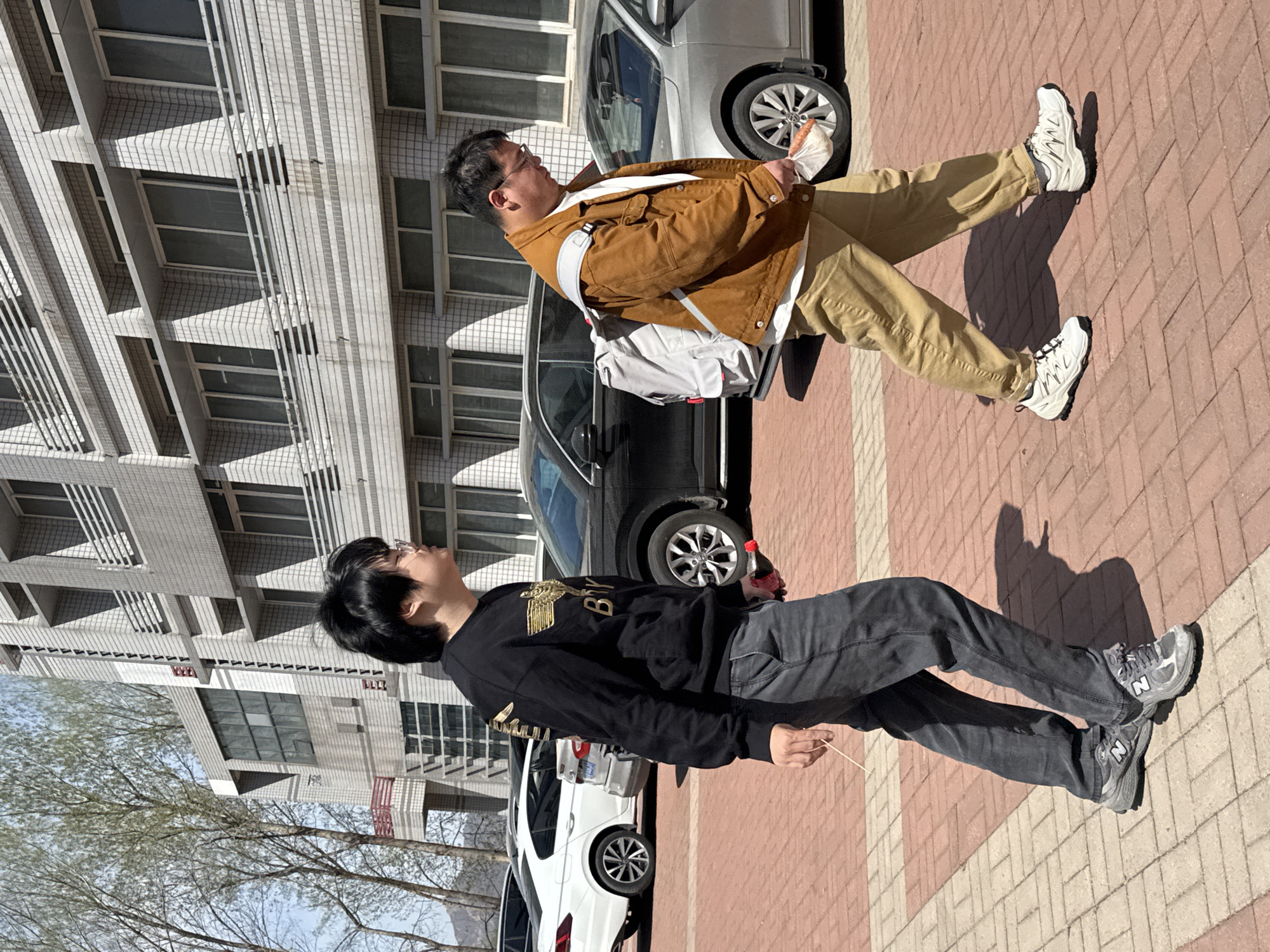 The image captures two men standing on a brick-paved sidewalk in an urban setting, looking upwards. The scene is brightly lit by the sun, casting distinct shadows on the ground, suggesting it is either mid-morning or late afternoon on a clear day.
The man on the right, appearing older, wears a brown button-up jacket over a white t-shirt, khaki pants, and white and black chunky sneakers. He carries a light grey sling bag across his body and holds a small, crinkled white bag, possibly containing food, in his left hand. He wears glasses and has short dark hair.
The man on the left, appearing younger, wears a black long-sleeved sweatshirt with elaborate gold embroidery, dark grey jeans, and grey New Balance sneakers with a visible "N" logo. He also wears glasses and has dark, neatly styled hair. In his right hand, he holds a thin wooden stick or skewer, and in his left, a small red-capped bottle, likely a beverage like Coca-Cola. His sweatshirt features gold text, with "BY" and part of an eagle-like emblem visible.
Behind the men, several cars are parked, including a silver SUV/van, a black SUV, and a white sedan. The background is dominated by a multi-story building with a light-colored, tiled facade and numerous windows, typical of residential or commercial architecture in China. Bare trees line the left side of the frame, indicating late winter or early spring. Both men have a neutral, observational expression as they gaze upwards.