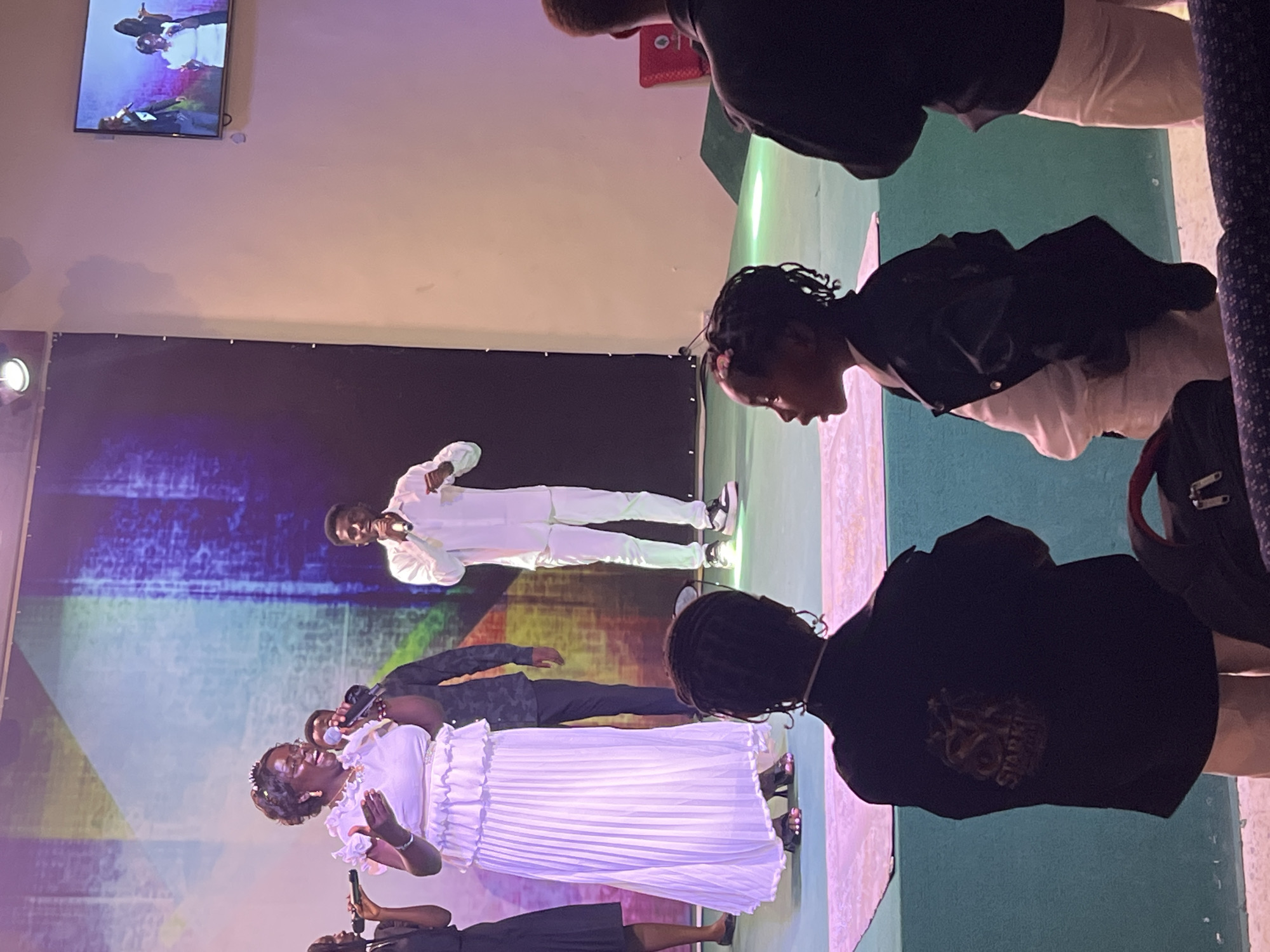 This image depicts a group of people on a stage in what appears to be a church or event hall in Gura Top, Nigeria. In the foreground, two children are seated, facing away from the camera, observing the performance. On the stage, a woman in a white, pleated dress and a man in a white suit are singing into microphones, accompanied by other individuals. The backdrop features a large screen displaying abstract, colorful imagery. A television screen mounted on the wall to the right shows a live feed of the performance. The environment appears to be indoors, with stage lighting illuminating the performers.