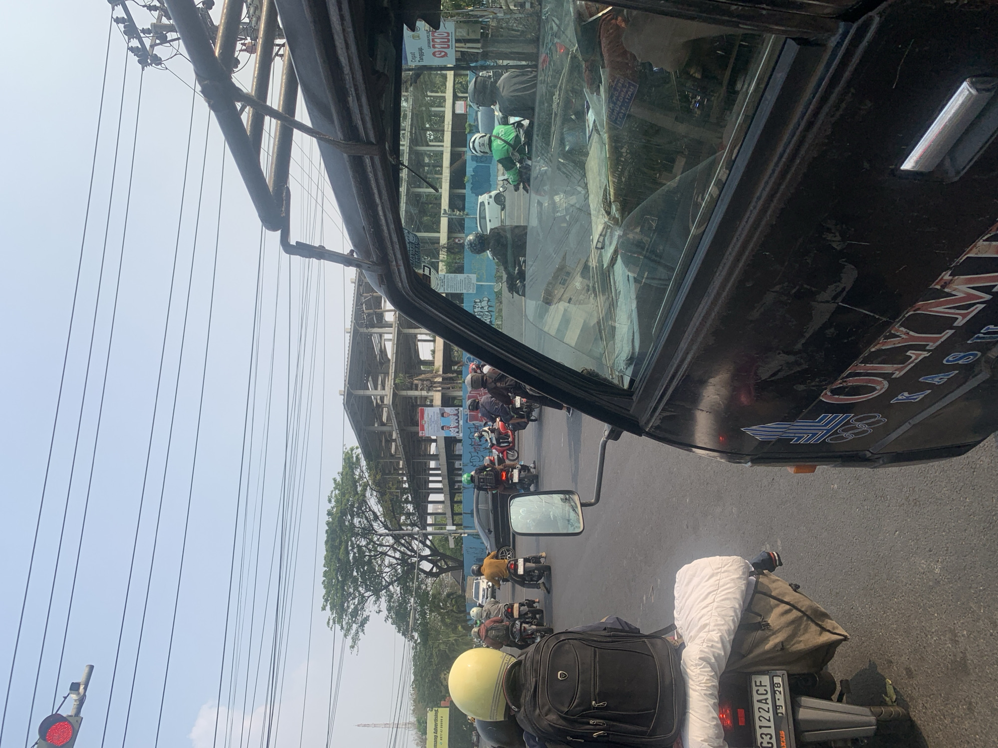 The image depicts a street scene in Palembang, Indonesia, likely during daytime traffic. In the foreground, a dark-colored vehicle, possibly a van or truck, is visible from the side, with part of its windshield and door showing. A logo with "OLYM" is partially visible on the side of this vehicle. To the left, a motorcycle with a yellow helmet on the rider is prominent. The motorcycle is carrying a white blanket or sheet. The license plate of this motorcycle reads "G3122ACF 09-28". The street is filled with other motorcycles and riders, suggesting moderate traffic congestion. In the background, a large, unfinished building structure is visible, along with trees and power lines crisscrossing the sky. The sky is clear and blue. There is a traffic light with a red signal illuminated in the upper left corner of the image. The overall environment appears to be an urban setting with ongoing construction.