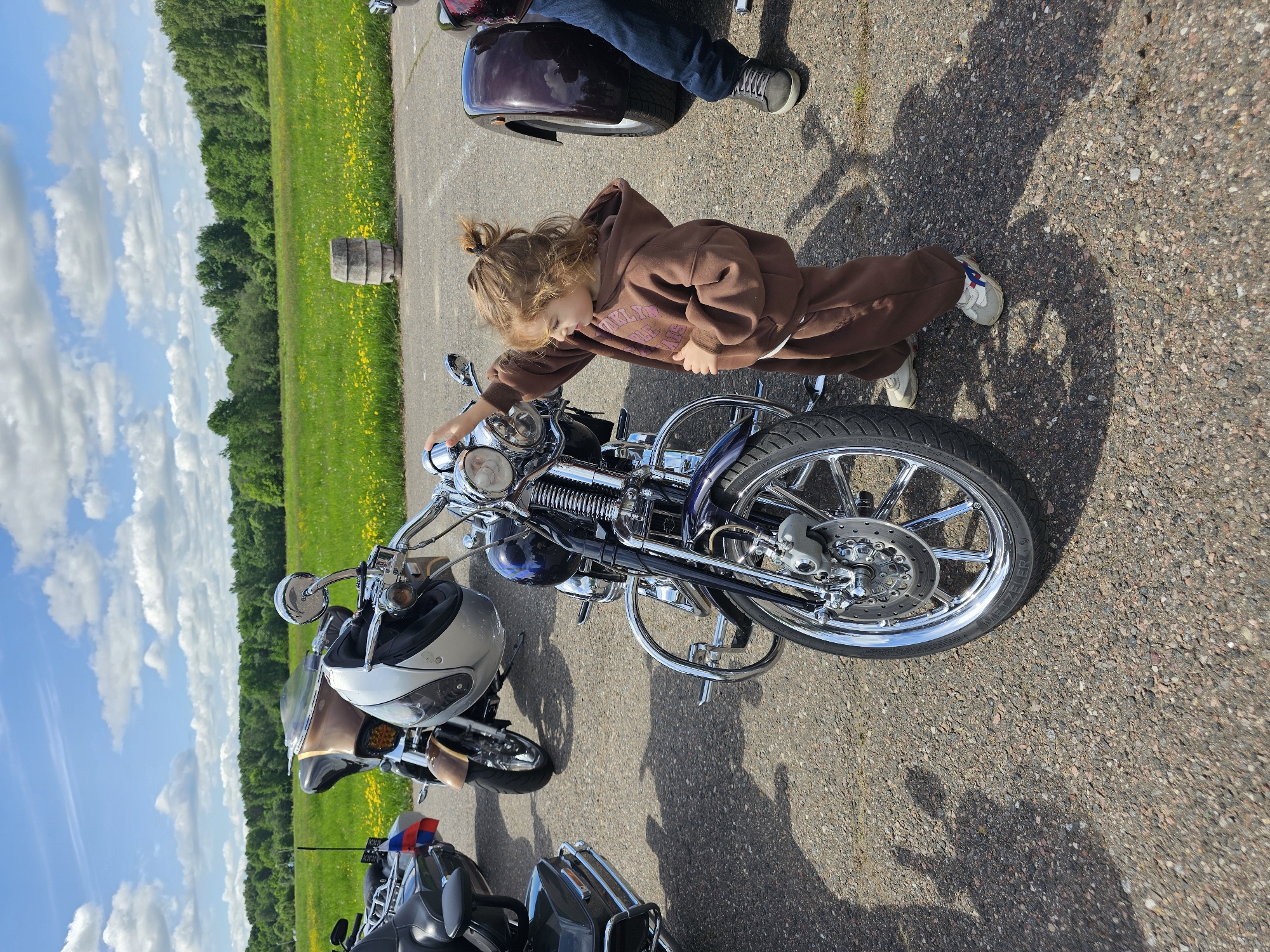 A young child wearing a brown hooded sweatshirt and pants is standing next to a motorcycle, seemingly interacting with the handlebars or controls. The motorcycle is chrome and has a blue gas tank. The scene appears to be in an open area with other motorcycles parked nearby. A field with yellow flowers and trees is visible in the background, under a sky with scattered clouds. The exact location within Borodinskogo Muzeya, Russia, is difficult to determine from the provided image alone, but it is likely an outdoor area near parking for visitors.