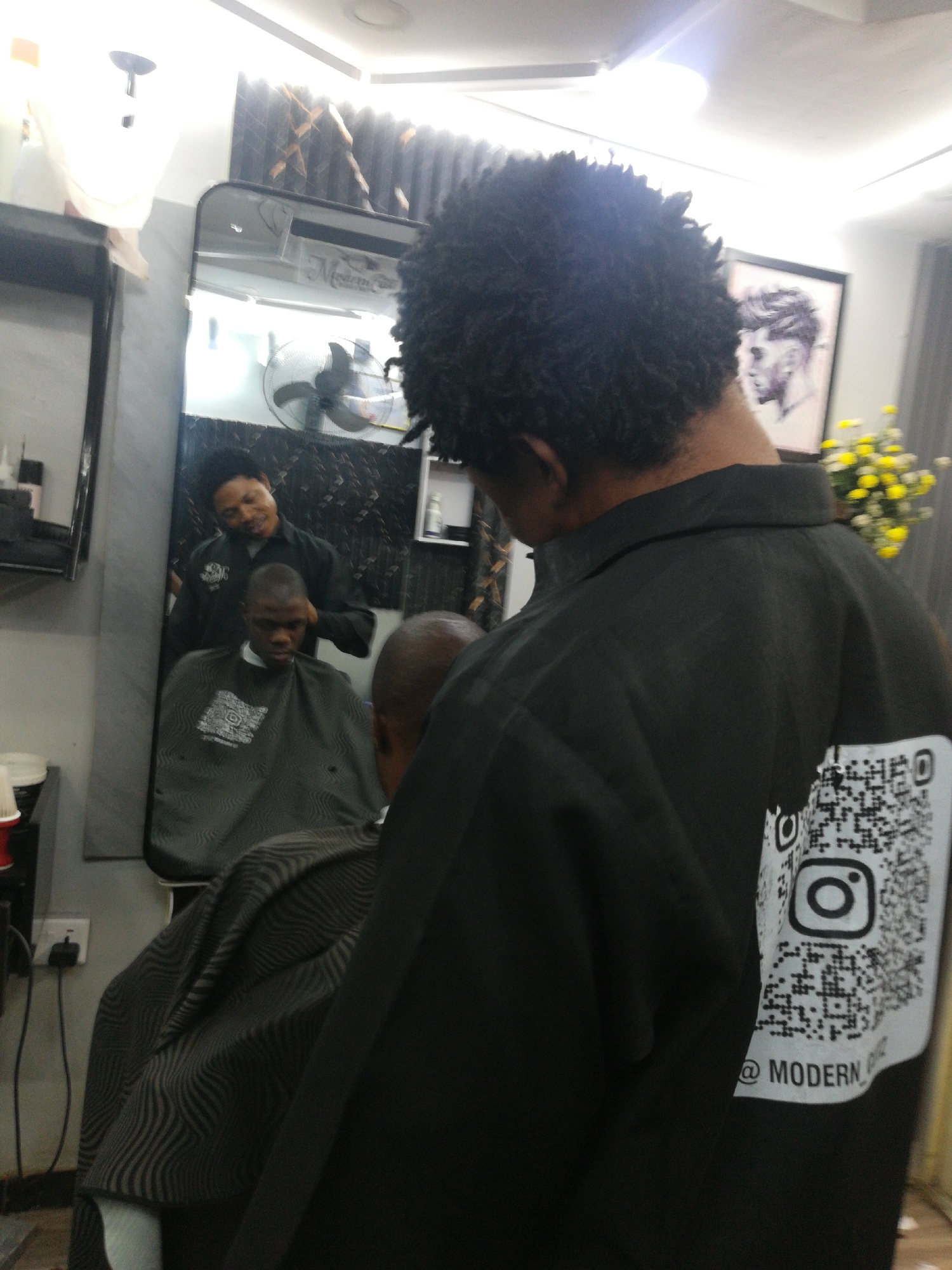 A medium shot captures a scene within a barbershop in Lagos, Nigeria. The barber, wearing a black jacket with a QR code and the word "MODERN" displayed on the back, stands close to a client who is seated and draped with a black and gray striped cape. In the background, a mirror reflects the barber interacting with the client. A fan is visible behind the reflected image. On the left side of the mirror, there's a black shelf, and to the right, a framed picture and yellow flowers. An electrical outlet is on the floor near a red and white container.