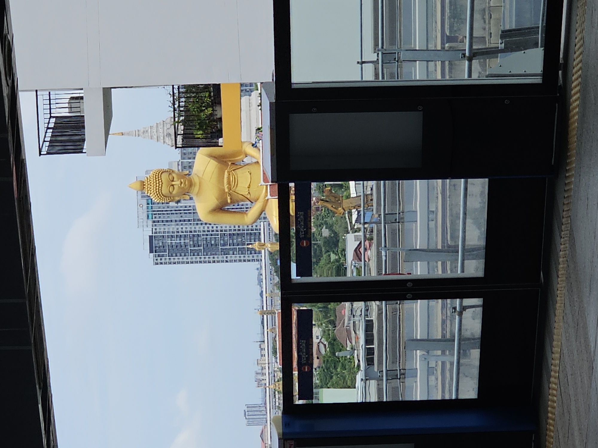 The image shows a golden Buddha statue on a raised platform, overlooking a cityscape. The statue is large and prominently featured in the center of the image, with its head and upper body visible. Behind the statue, a white building with balconies and a distinctive stupa-like structure can be seen. The cityscape in the background includes tall, modern buildings and smaller residential structures, all set against a cloudy sky. In the foreground, the image captures the glass doors and platform of a train station, with railings and tracks visible. The overall scene suggests an urban environment, likely in Bangkok, Thailand, where such large religious statues are often integrated into modern architecture.