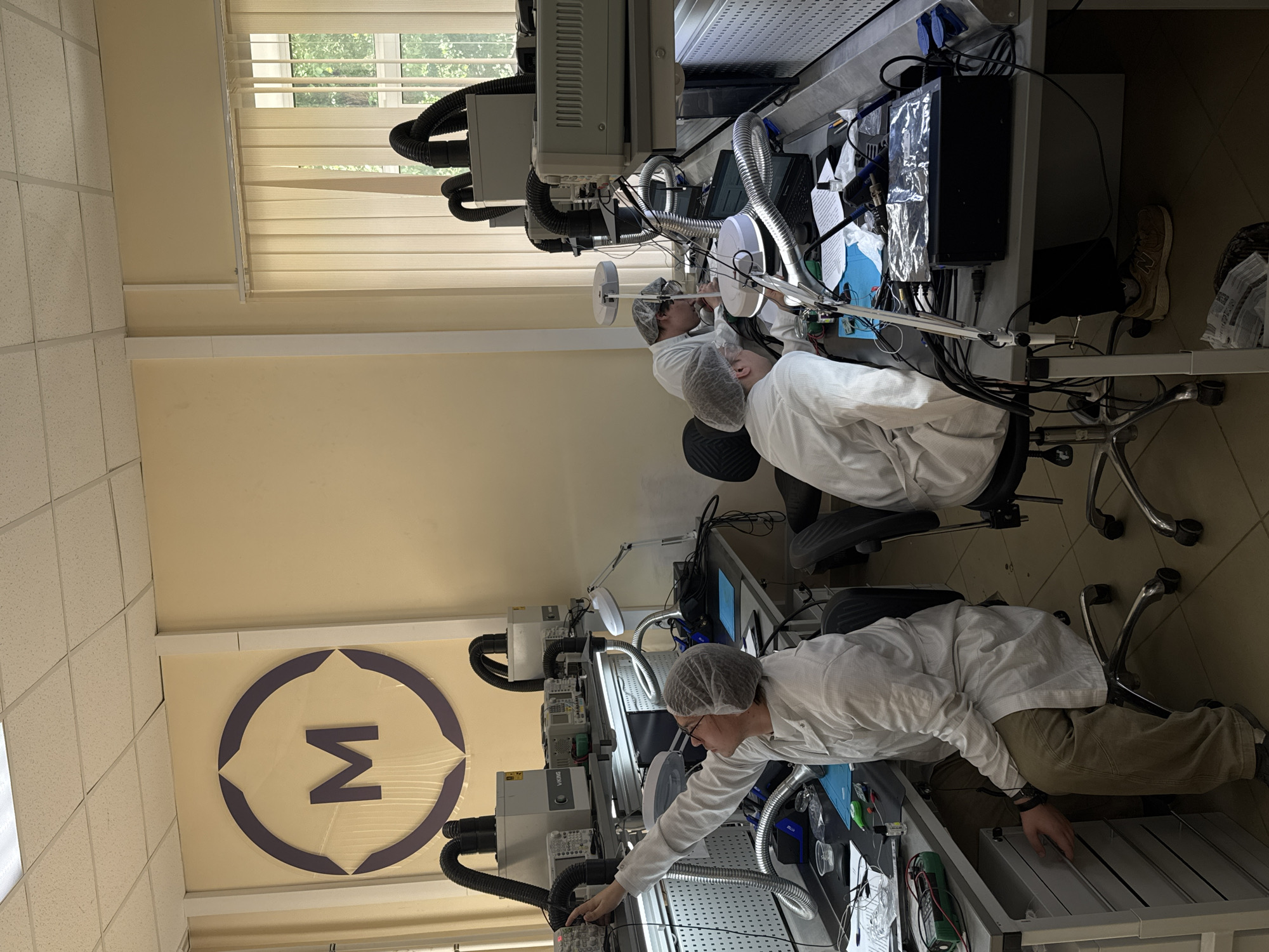 In an indoor laboratory in Perm, Russia, three individuals wearing white lab coats and hairnets are working at a series of interconnected workstations. Each station includes various electronic devices, including scopes and power supplies connected with numerous wires and tubes. The walls of the lab are painted a light yellowish color, and a logo depicting a stylized sigma symbol is visible. Natural light enters the room through a window with closed blinds. The individuals appear to be focused on their work, manipulating equipment and examining components.