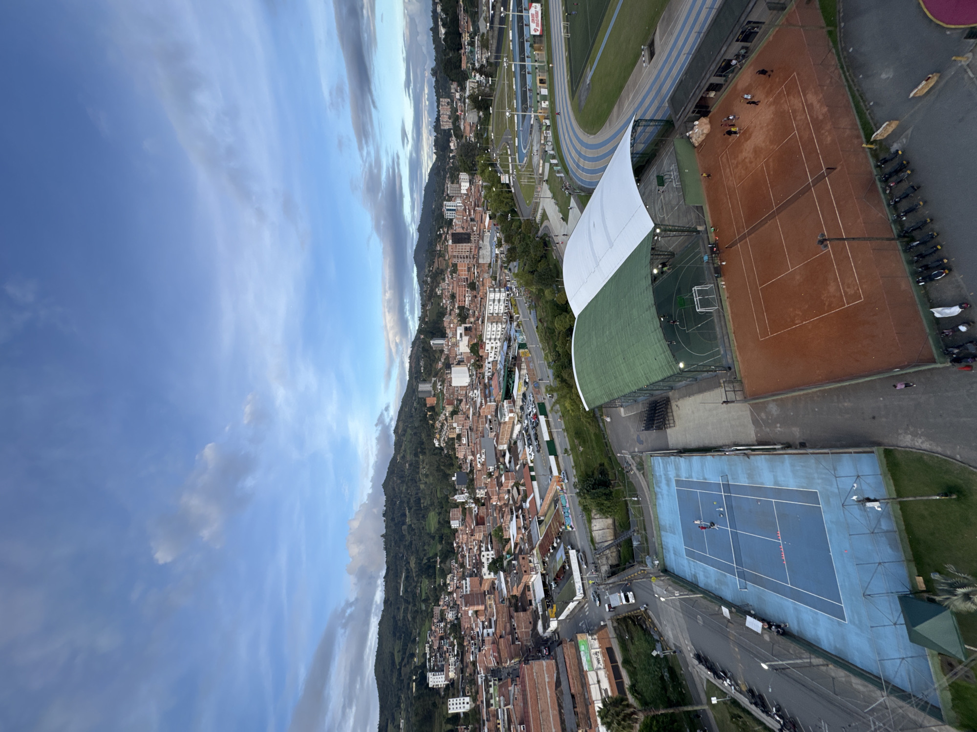An aerial, high-angle, wide shot captures a panoramic view of the city of Guarne, Colombia, at dusk. The sky is a gradient of blue with scattered, wispy clouds and some darker, more substantial clouds closer to the horizon. In the foreground, there are two tennis courts; one is blue with white lines, and the other is clay with white lines. Several people are visible on both courts, engaged in activities. To the right of the tennis courts is a running track with blue and white lanes, adjacent to a soccer field. Beyond the sports facilities, a dense urban area with red-tiled roofs and brick buildings stretches out, nestled against lush green hills. Various vehicles, including cars and motorcycles, are parked along the streets and in parking lots. The overall scene depicts a lively community with a focus on sports and recreation, set against a backdrop of residential housing and natural landscape.