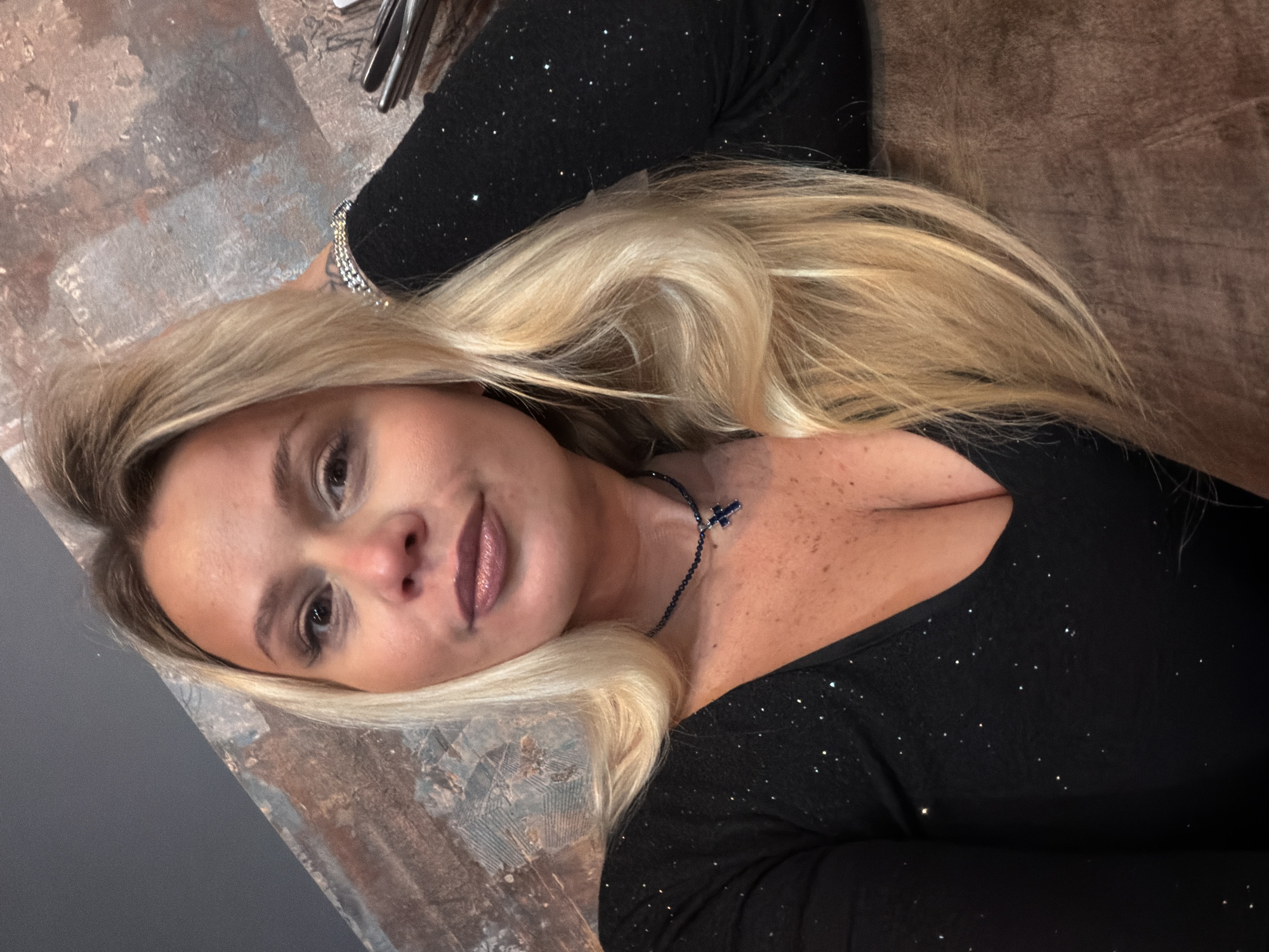 An indoor portrait shows a blonde woman looking directly at the camera. She has long, light blonde hair styled over her shoulders and a visible left hand resting behind her head, slightly touching her hair. Her eyes are dark, and she wears subtle makeup including eyeliner and lip gloss. She is dressed in a black, long-sleeved V-neck top adorned with scattered sparkling elements. Around her neck is a dark beaded necklace with a small blue cross pendant. A multi-row, silver-toned bracelet is visible on her right wrist. The background features a wall with a textured, abstract pattern combining dark gray, brown, and reddish-brown hues. To the right, a dark, possibly upholstered surface and metallic objects resembling cutlery are partially visible, suggesting a cafe or restaurant setting. The specific location within the city of Sarapul, Russia, is not identifiable from the visual content.