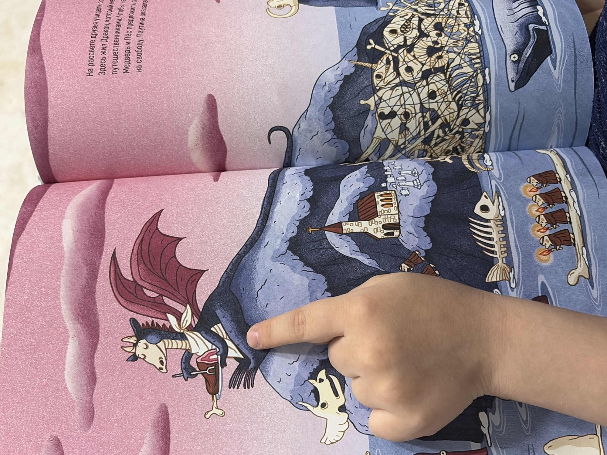 The image shows a child's hand pointing at an open children's book, likely in Chaikovsky, Russia. The book displays colorful, fantastical illustrations and Russian text.
On the left page, against a pink, textured background, a dark, winged, dragon-like creature with a white bib is perched on a rocky, dark blue formation. It holds a fork and knife, poised to eat a piece of meat on a stand.
The right page continues the rocky, dark blue landscape, extending into blue water or sky. In the upper right, a large shark-like fish swims next to a dense pile of human skulls and bones, some appearing to be bound. Below this, a small village or castle with multiple spires is depicted on the cliff face. Further along the rocky edge and water, a skeletal fish is visible, followed by several small, robed figures holding candles or torches, arranged in a line.
Visible Russian text on the left page reads in fragments: "At dawn, the friends saw...", "Here lived a Dragon, who did not...", "travelers. In order not to...", "Bear and Dog suggested...", and "to freedom. The cobweb turned out to be...". This suggests a narrative involving a dragon, companions, and an adventure or escape. The scene combines whimsical elements with some macabre details within a fantastical setting.