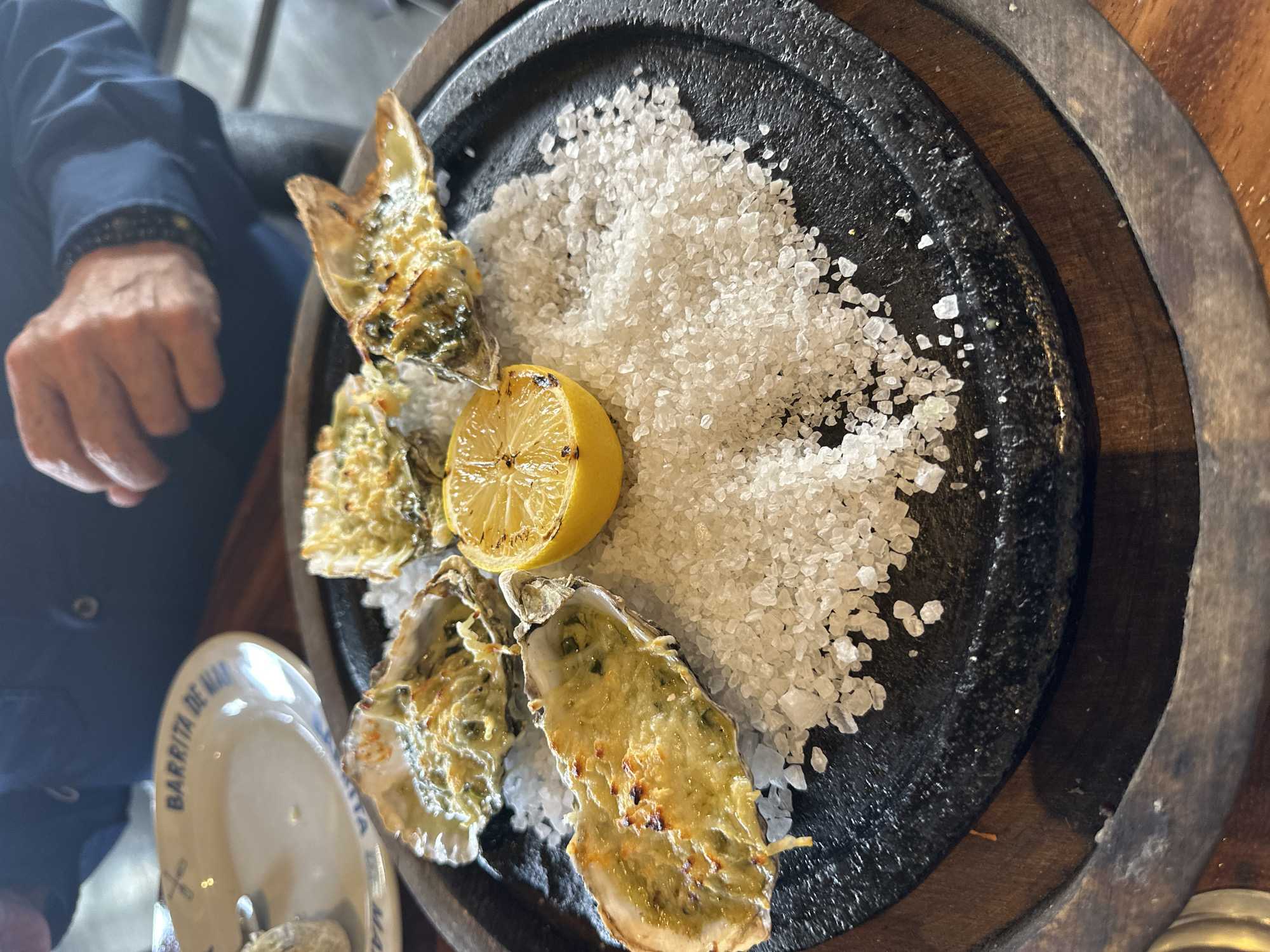 The image shows a plate of grilled oysters served on a bed of coarse sea salt with a grilled lemon half. The oysters have a melted cheese or creamy sauce topping. The plate appears to be made of black stone and is set on a wooden surface. Part of a plate with the inscription "BARRITA DE MAR" is visible in the lower left corner. A person's hand and a blue shirt sleeve are visible on the left side of the frame. The image was taken at a restaurant in Huixquilucan de Degollado, Mexico, specifically the establishment "BARRITA DE MAR".