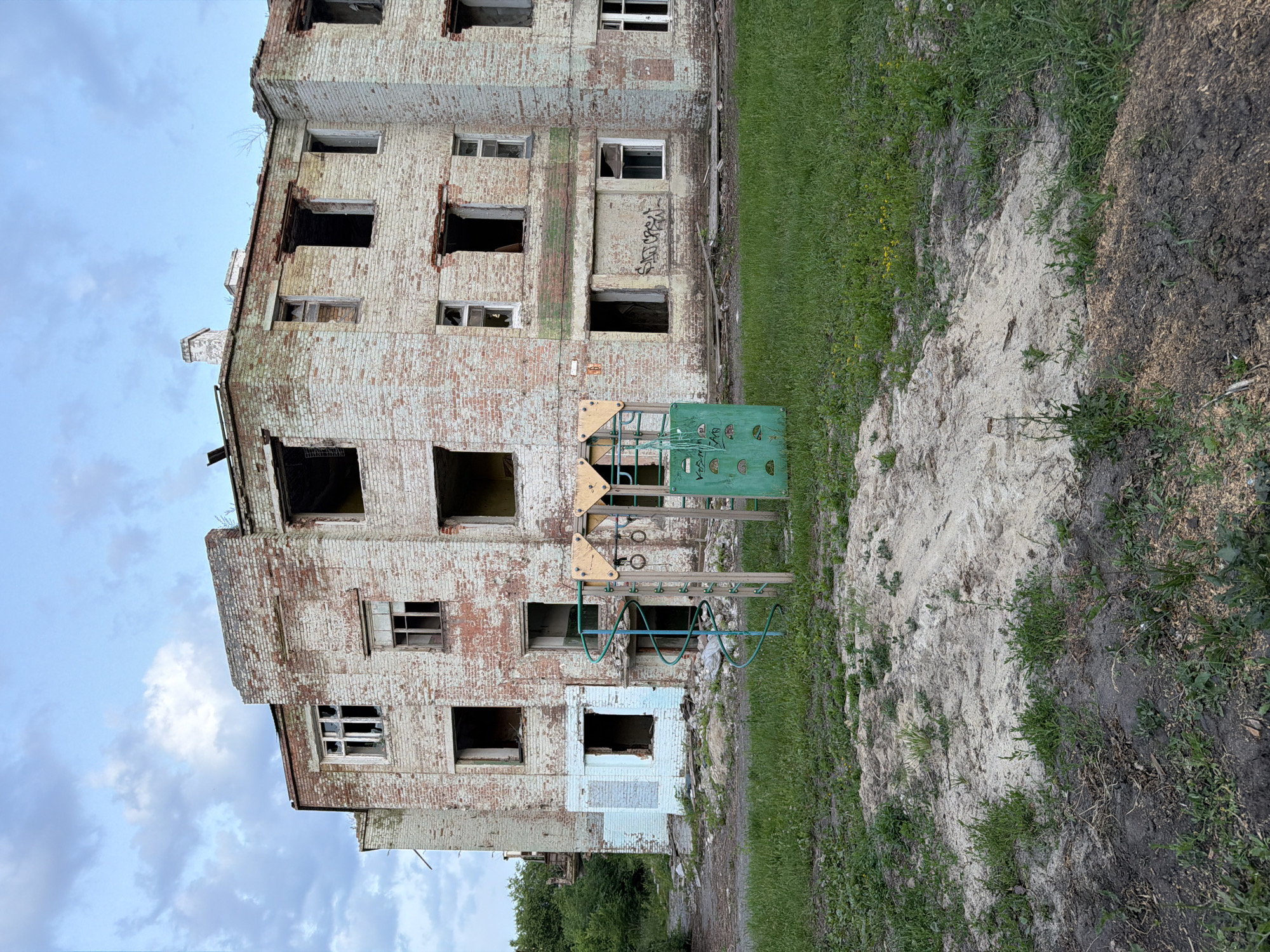 The image shows the exterior of a dilapidated, multi-story brick building with numerous broken windows and signs of decay. The building appears to be abandoned, with peeling paint and visible structural damage, particularly at the upper levels.
In front of the building, there is a weathered playground structure, painted green, with metal rings and some wooden elements. The playground is situated on a patch of dirt and sand, with overgrown grass and weeds surrounding it. A pile of sandy soil and dark earth is visible to the right of the playground. The building and playground are set against a backdrop of a cloudy sky.
There is some graffiti visible on the building, particularly on a section of the wall near the playground and on a doorway. The overall scene evokes a sense of abandonment and neglect, possibly in an urban or semi-urban environment given the presence of the building and playground. The time of day appears to be late afternoon or early evening, judging by the quality of light and the cloud cover. The location context is Semiluki, Russia.