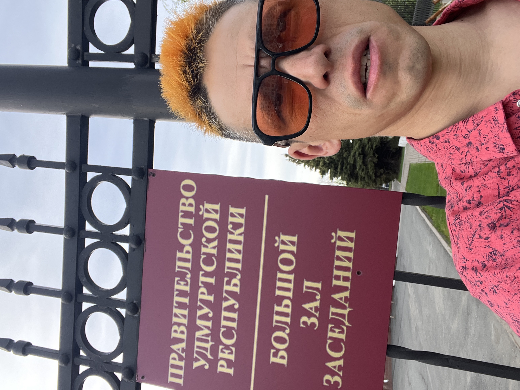 The image shows a man wearing sunglasses and a pink shirt with a floral pattern standing in front of a sign that reads "Government of the Udmurt Republic, Large Meeting Hall" in Russian. The sign is attached to a black metal fence with circular patterns. Behind the fence, trees and a gray walkway are visible. The location is Izhevsk, Russia, based on the text on the sign. The man is likely taking a selfie.