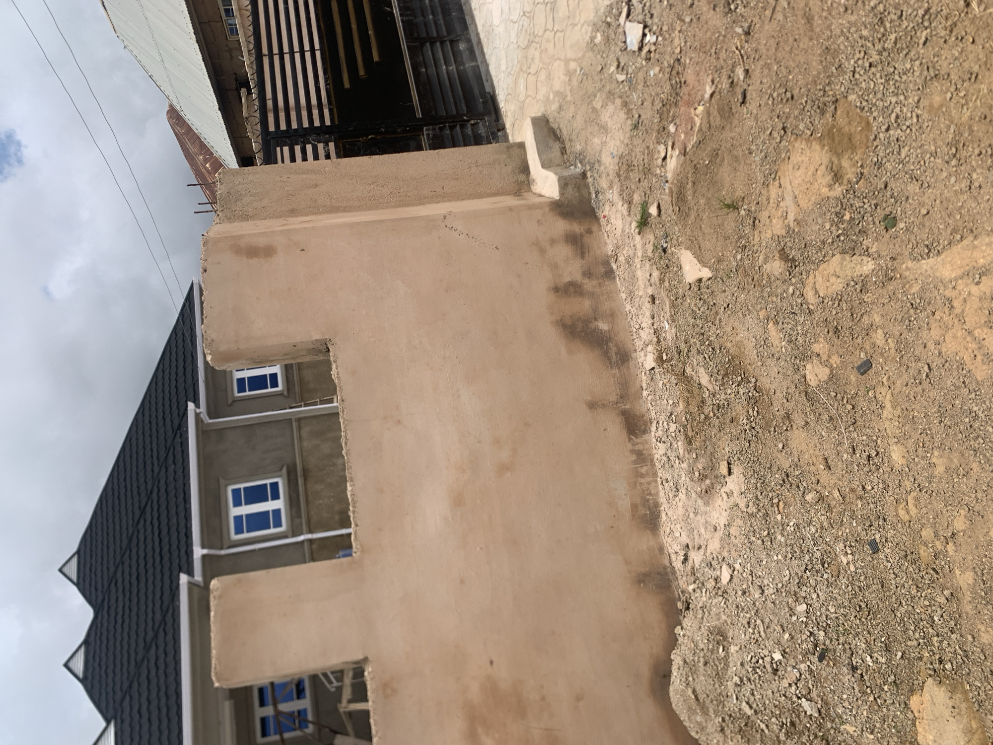 The image shows a construction site with a partially built wall in the foreground. Behind the wall, a house with a dark, corrugated roof and blue-tinted windows is visible. To the right, a black metal gate and a section of a corrugated metal roof are present. The ground is covered with loose gravel and dirt, indicating ongoing construction work. The sky is overcast with clouds. The scene depicts a typical residential construction project in the city of Jos, Nigeria.