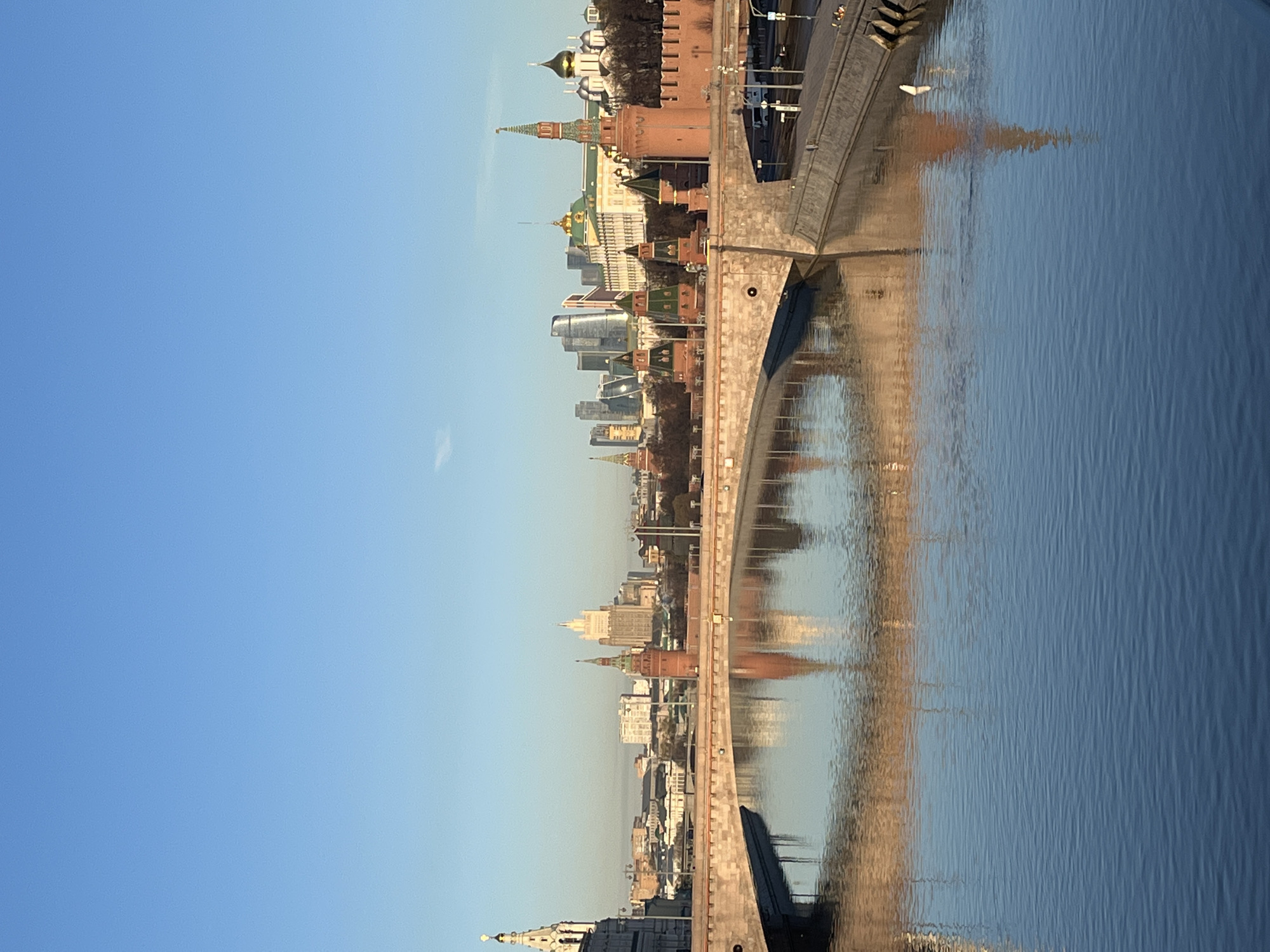 The image depicts a panoramic view of the Moscow Kremlin and its reflection on the Moskva River in Moscow, Russia. A bridge crosses the river in the foreground. The Kremlin's towers, cathedrals, and walls are visible, along with modern buildings of Moscow. The sky is blue. The water of the river reflects the buildings and the bridge.