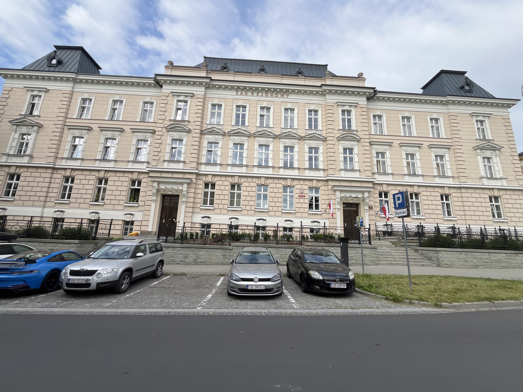The image shows a pale pink, three-story building with a dark gray mansard roof and black dormer windows. The building has many windows with white trim and shutters. Above the central windows, the word "SĄD REJONOWY" is inscribed. There are two flags, one red and white Polish flag, displayed on either side of the central entrance.
In front of the building is a parking lot paved with cobblestones. Several cars are parked there: a blue sports car, a silver SUV, a silver Volkswagen Golf, and a black hatchback. A gray parking sign with a blue 'P' and Polish text "Koniec" meaning "End" is visible. The parking lot is bordered by a low stone wall and a black metal fence. Steps lead up to the building's entrance, and grass grows in front of the stone wall. The sky is partly cloudy.