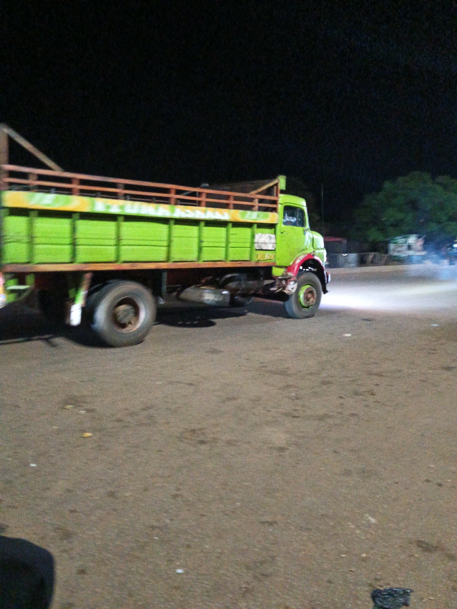An unidentifiable truck is shown at night in an outdoor location. The truck is bright green and appears to be a flatbed truck with wooden railings along the sides of the cargo area. The truck is in motion, as indicated by the blur on the tires and the smoky exhaust coming from the rear. The scene is dimly lit, suggesting it is nighttime. The ground is paved with asphalt, and there are some trees and buildings in the background.
