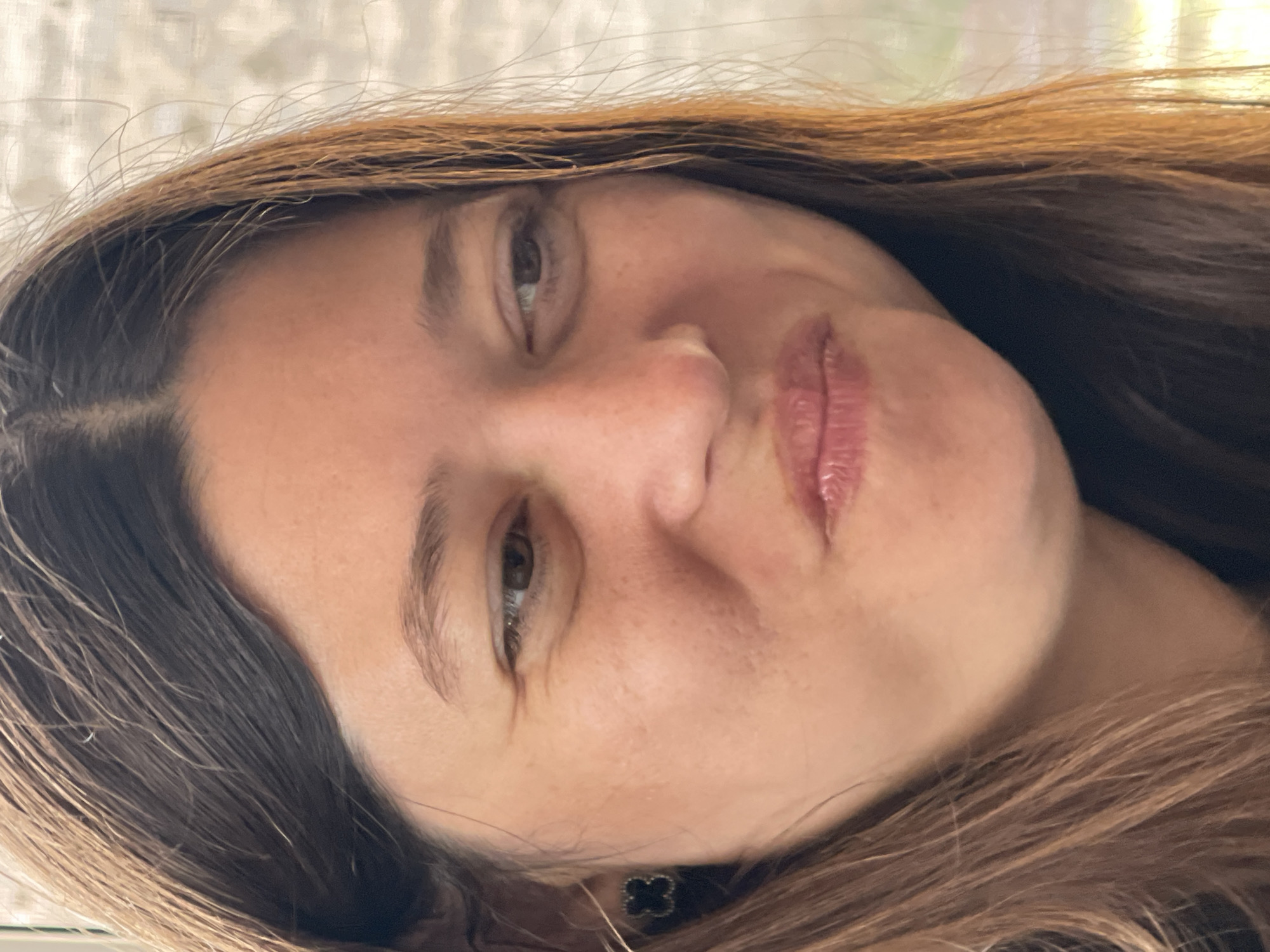 A close-up, slightly angled headshot shows a woman with brown hair and light skin. Her lips are pursed. She has brown eyes and visible freckles. A necklace with a black, floral-shaped charm is visible at the base of her neck. The background is a light, blurred pattern. The location and any specific forensic details are not discernible from the image.