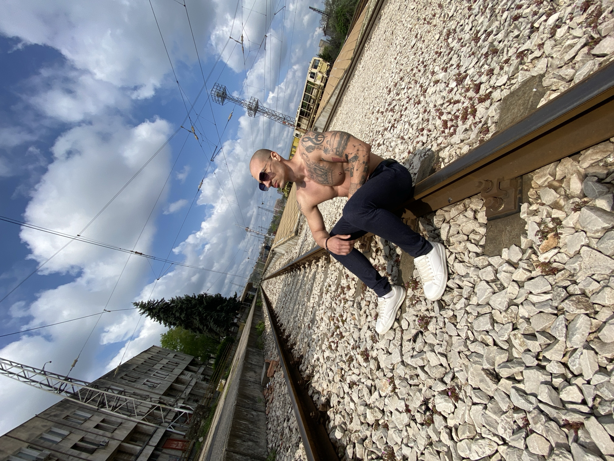 A bald, light-skinned man with sunglasses and extensive tattoos on his upper body is sitting on a railway track. He is wearing dark pants and white sneakers. The railway is set on a bed of light-colored stones. Overhead power lines are visible against a cloudy blue sky. A building and trees are visible in the background. The scene appears to be in an urban environment, near Pleven, Bulgaria, based on the surrounding architecture and infrastructure. A train carriage is visible along the tracks in the background.